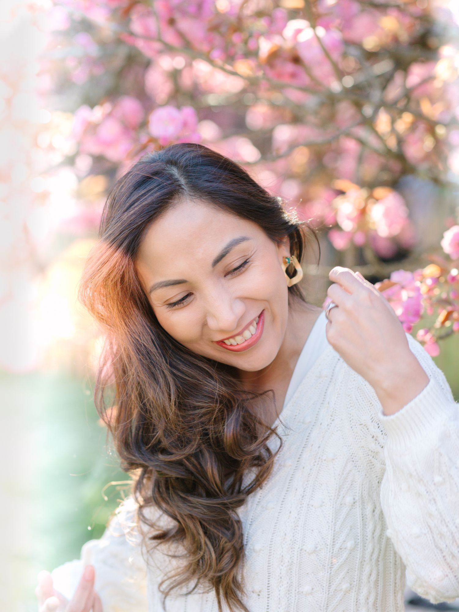 Smiling woman with long dark hair in white sweater posing outdoors near pink cherry blossom trees in spring.