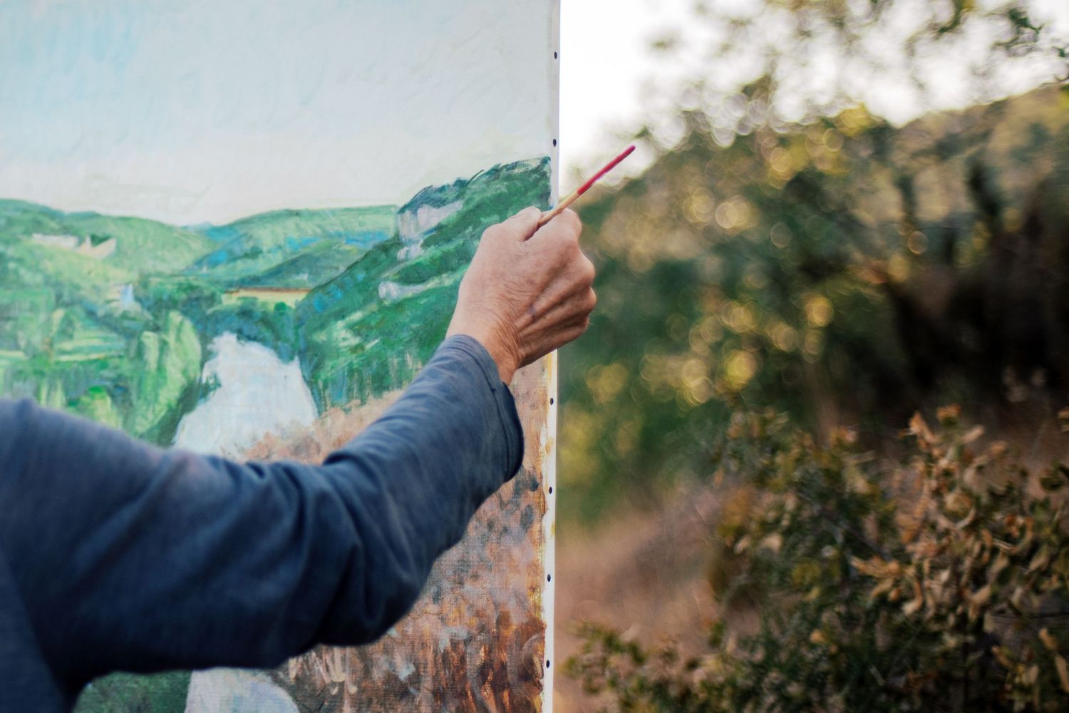 Artist painting a landscape on canvas outdoors, with a real green valley and river visible beside the artwork.