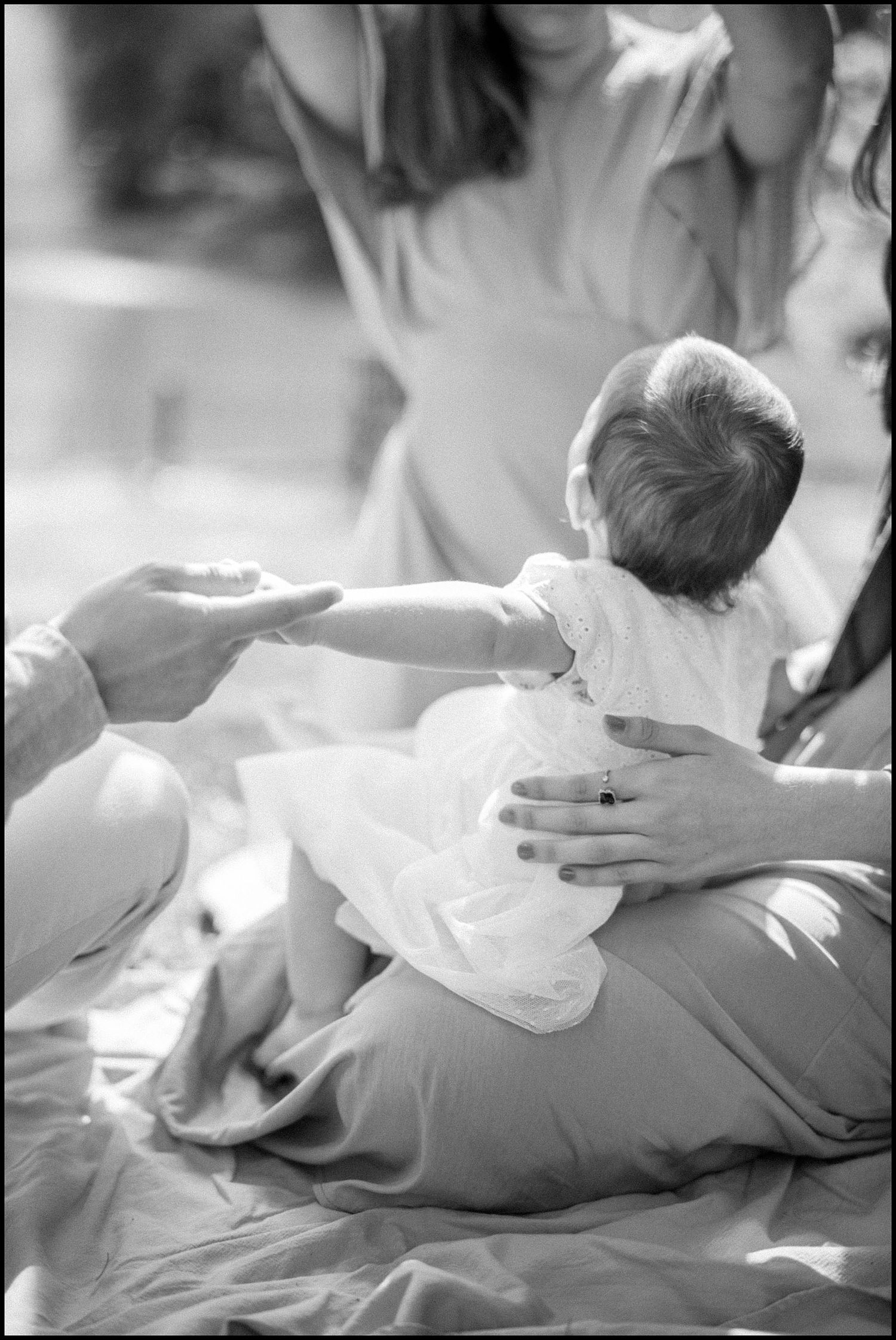 Black and white photo of a baby in white dress reaching out to an adult while being held by another person.