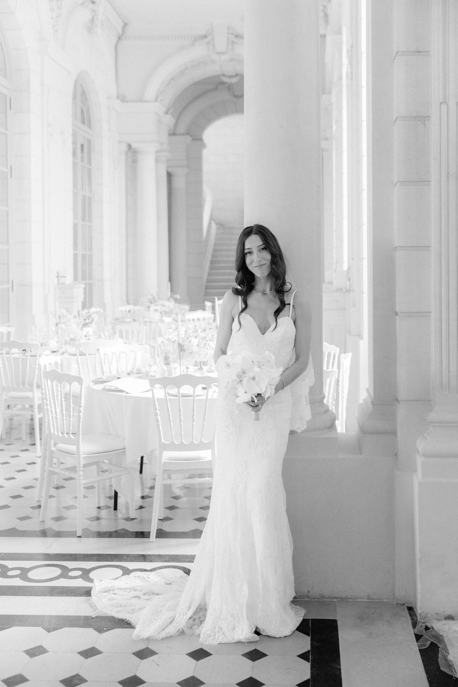 Elegant bride in white lace gown holding bouquet stands in grand hall with set reception tables, black and white photo.
