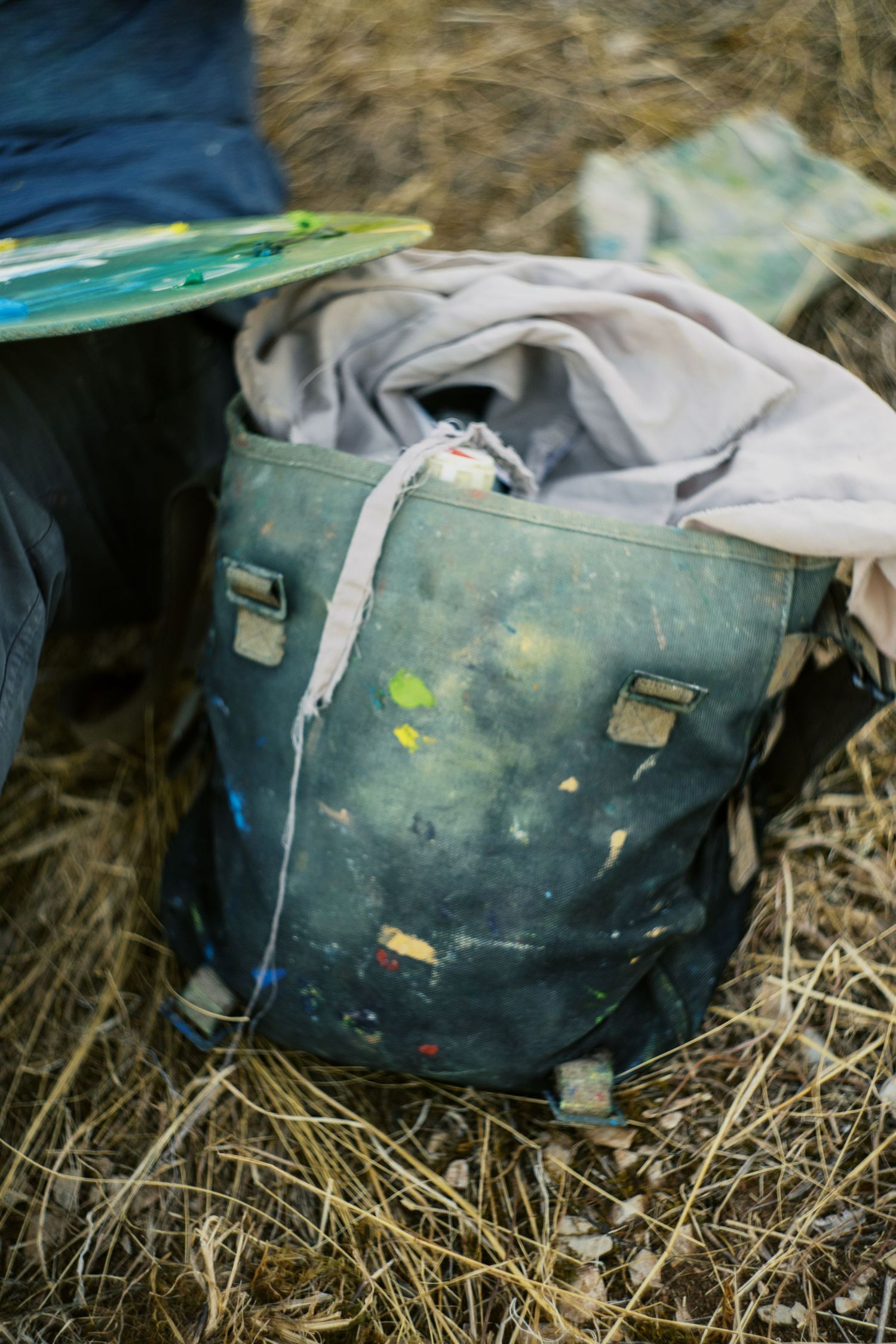 A paint-splattered dark green canvas backpack sitting on dry grass outdoors near an artist's palette.