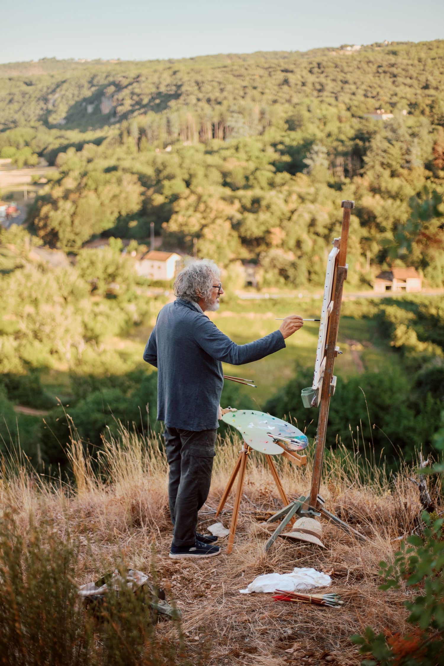 Elderly artist painting on easel outdoors on hillside with lush green forest landscape in background during golden hour.