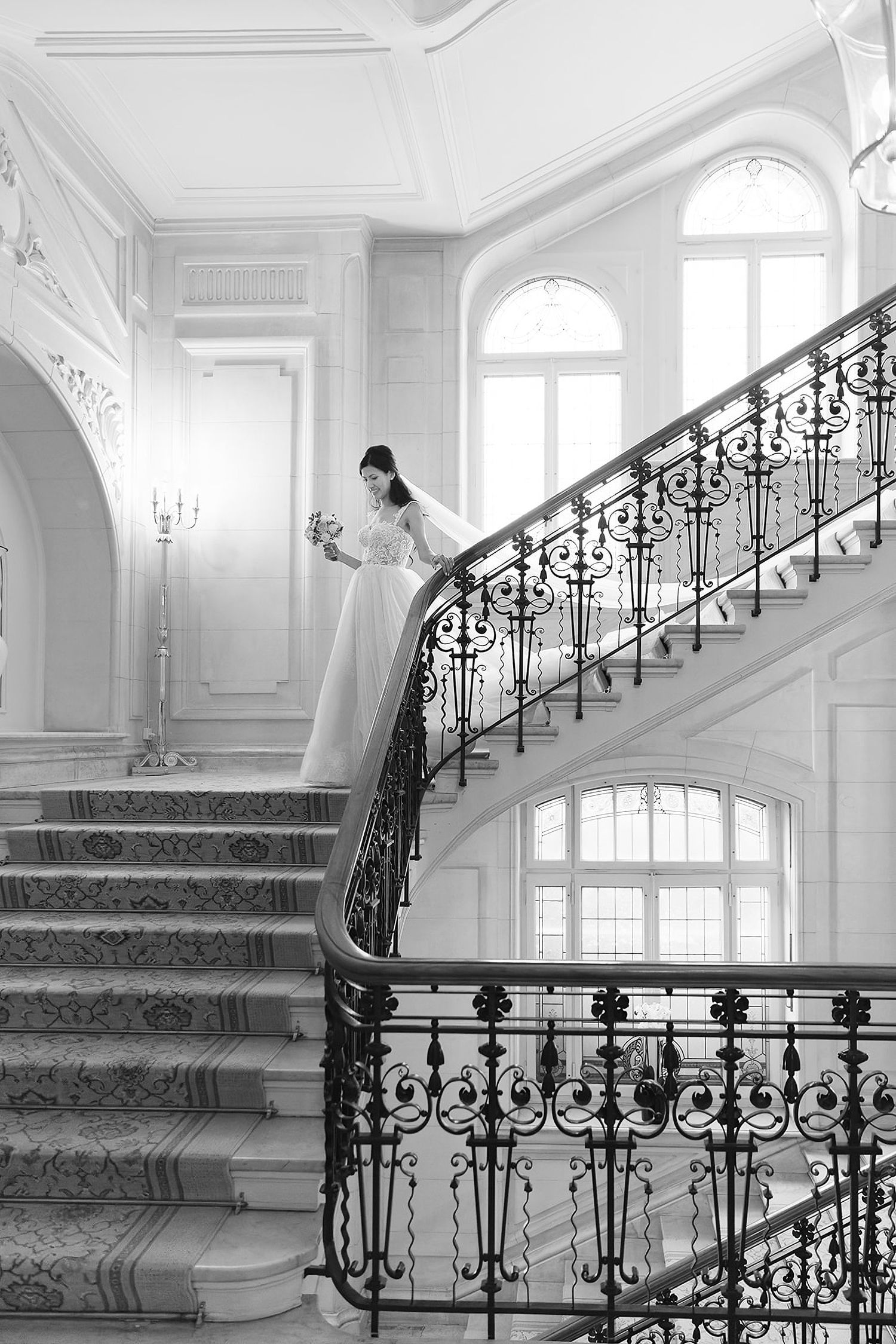 Bride in white gown descending ornate wrought iron staircase in elegant black and white wedding photo.