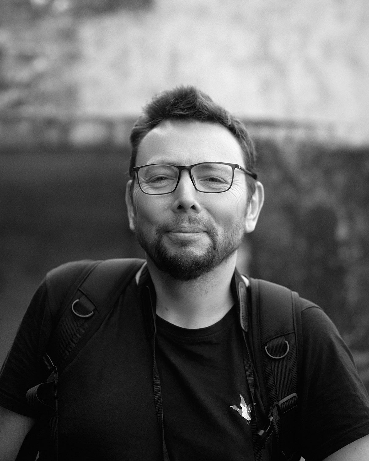 Black and white portrait of a person wearing glasses and a black t-shirt with a backpack against a blurred background.