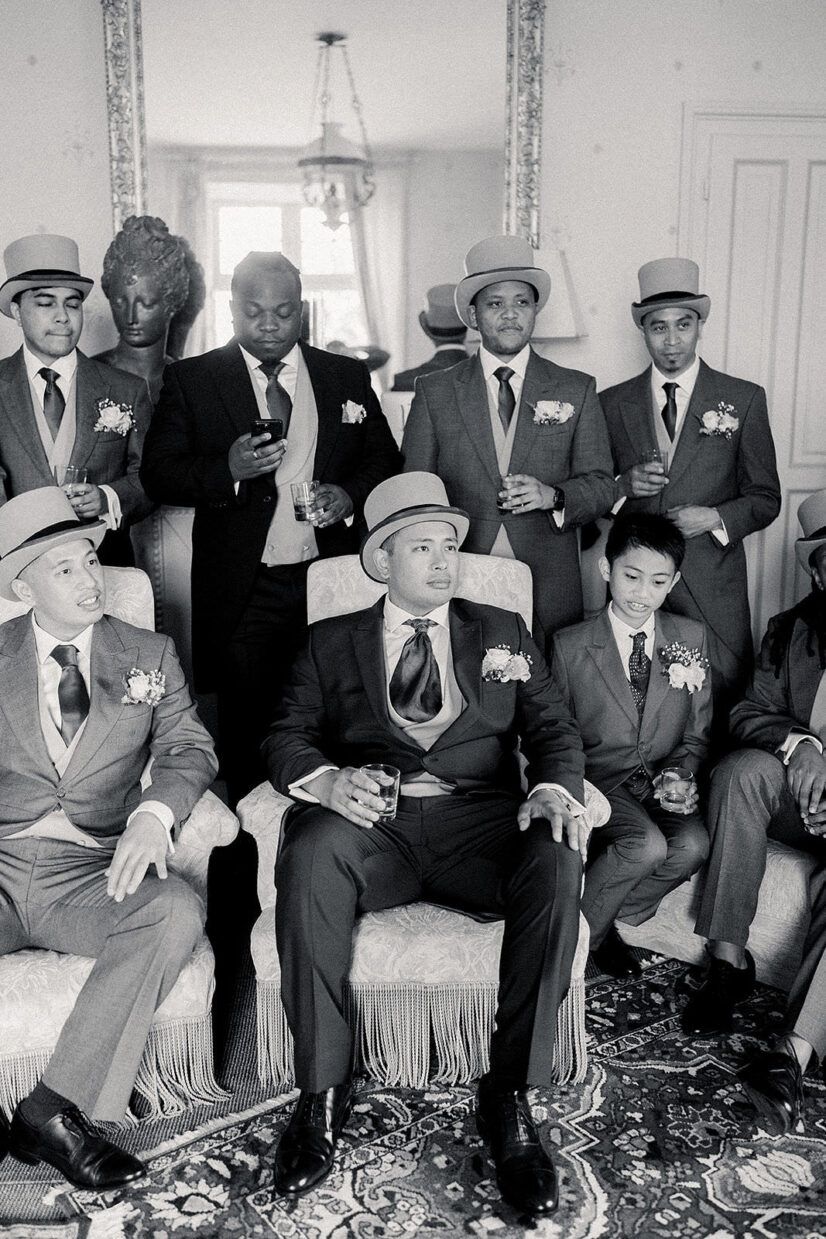 Black and white photo of groomsmen in suits and hats posing together in an elegant room before a wedding ceremony.