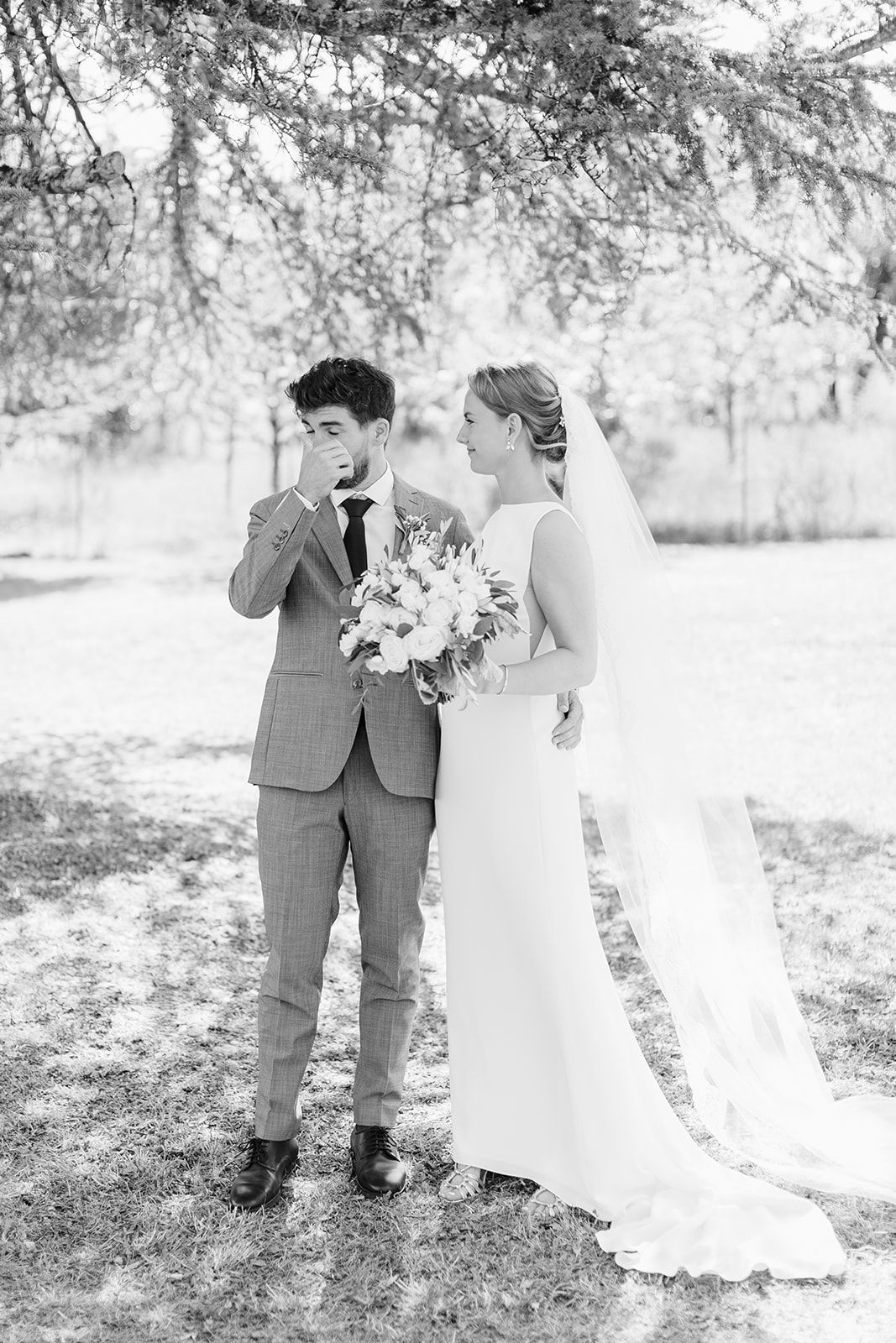 Black and white photo of a bride holding bouquet and groom in suit standing under a tree on their wedding day.