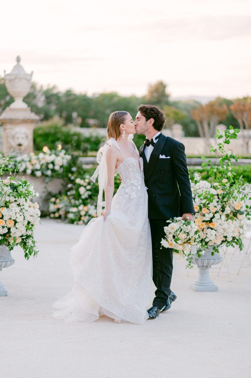 Bride and groom share a romantic kiss at outdoor wedding ceremony surrounded by elegant floral arrangements in stone urns.