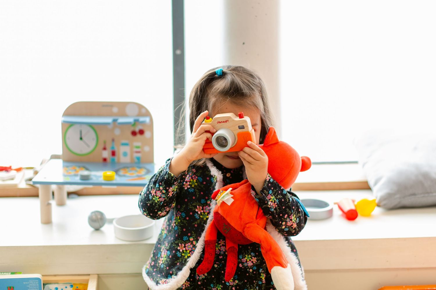 Young girl holding a stuffed fox toy while looking through a toy camera in a bright playroom.