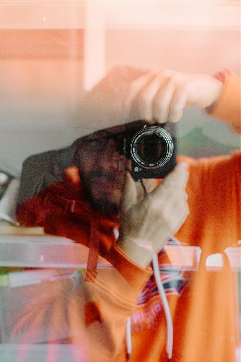 Reflet artistique d’une personne prenant une photo à travers une vitre, dans des tons orange et bleu-vert.