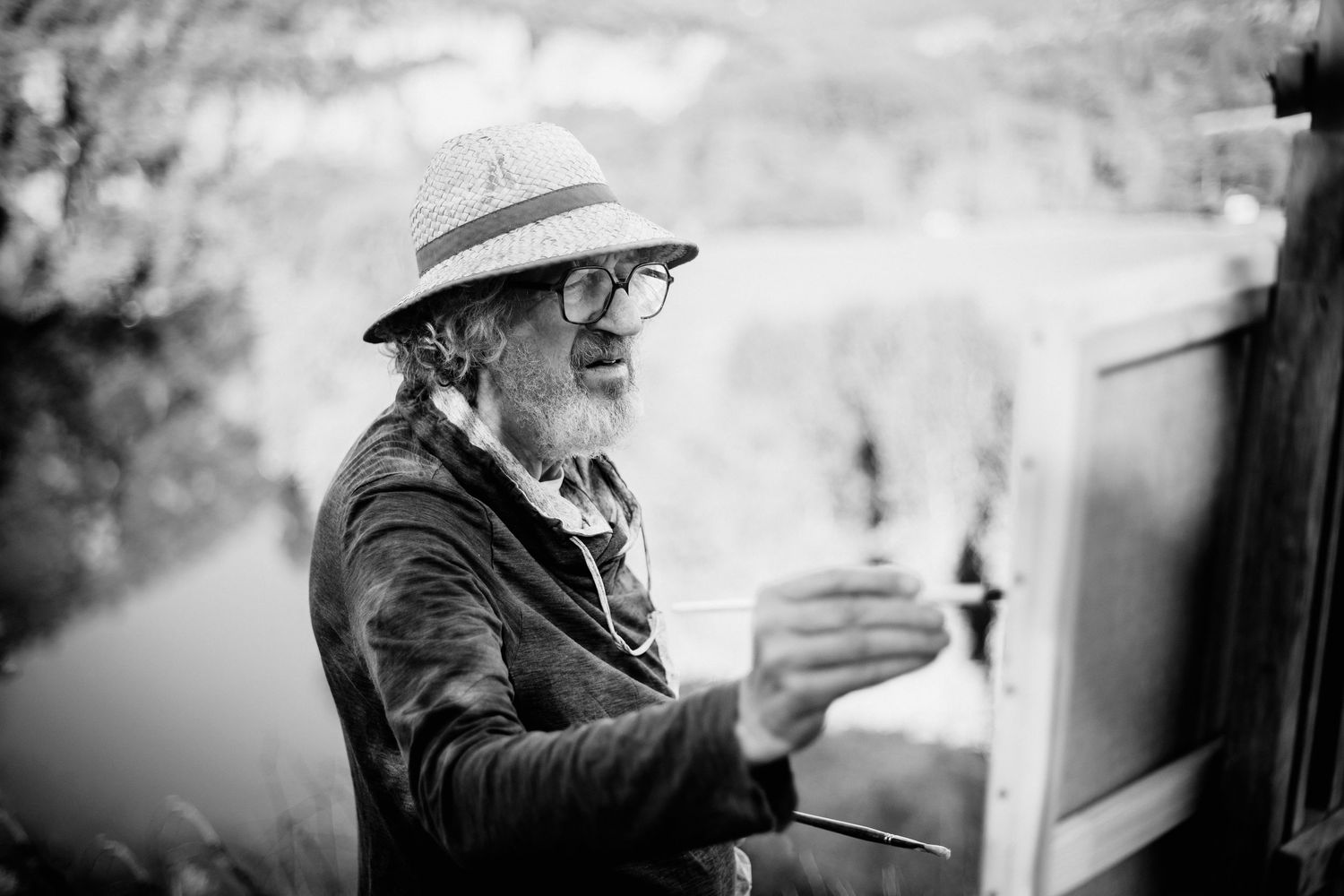 Elderly male artist painting outdoors on canvas at an easel, wearing a hat and glasses, black and white photo.