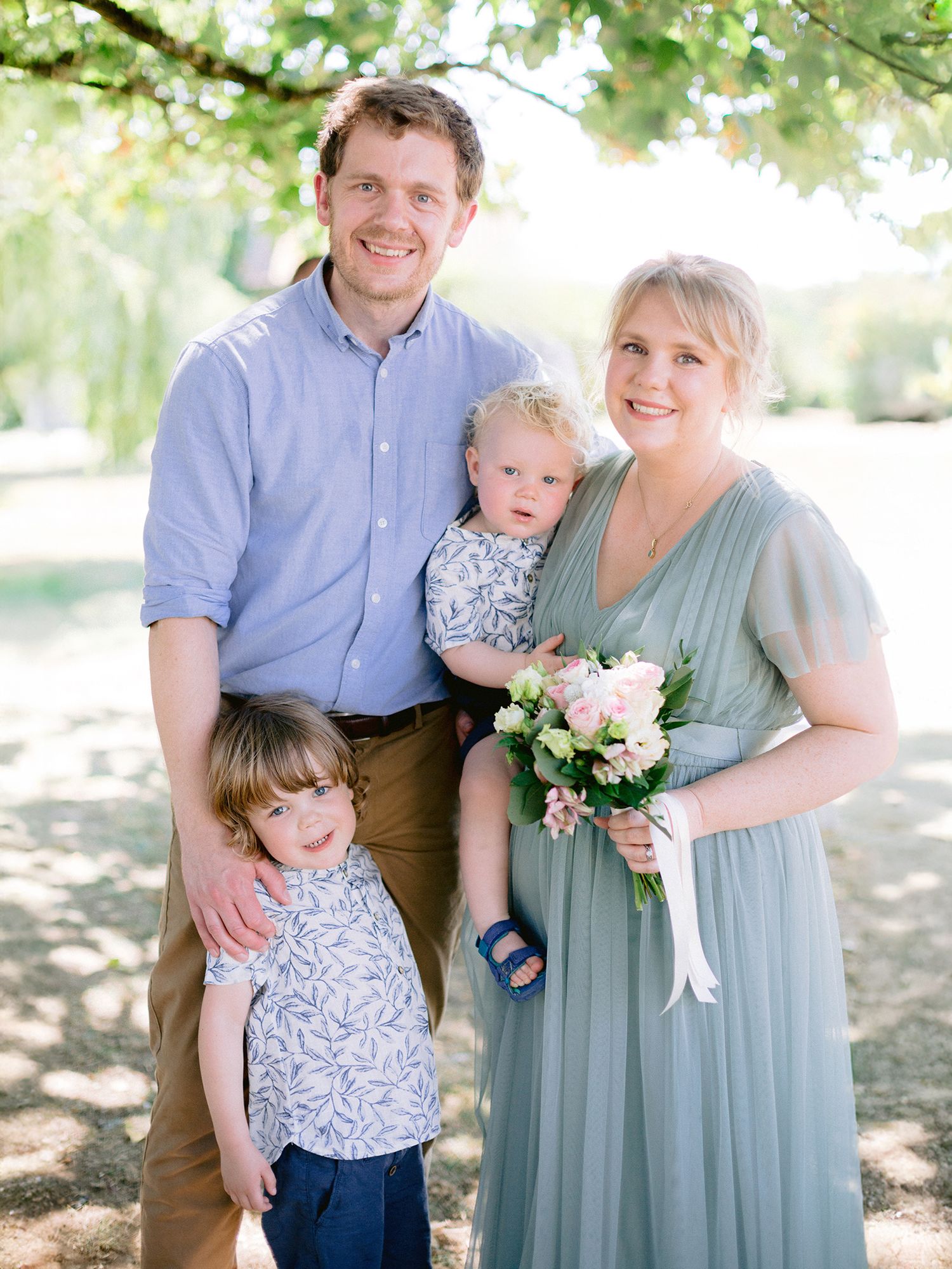 A family of four poses together outdoors under dappled sunlight, with the person on the right holding a floral bouquet.