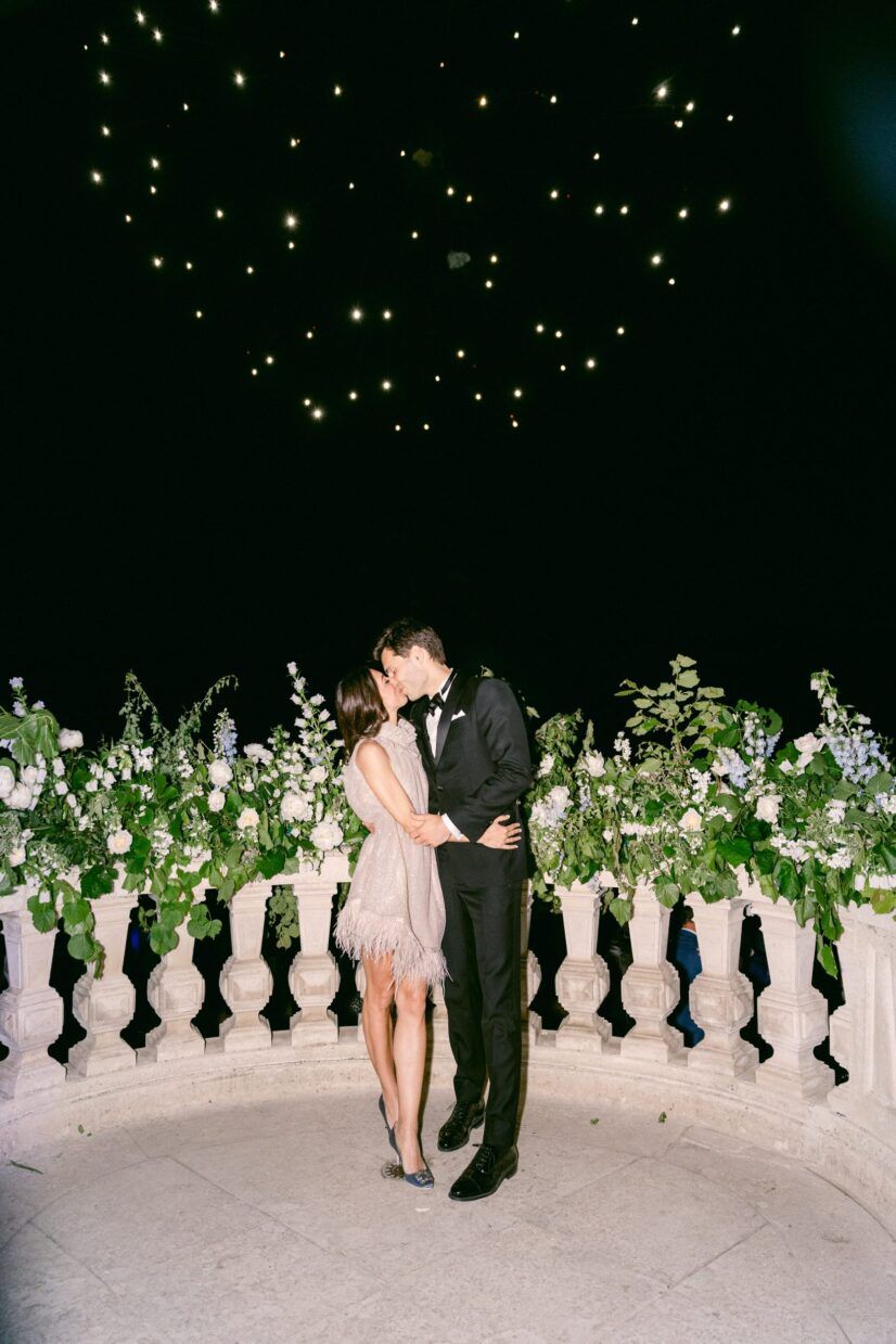 Couple kissing on a moonlit balcony with white floral arrangements and twinkling lights at a romantic wedding.