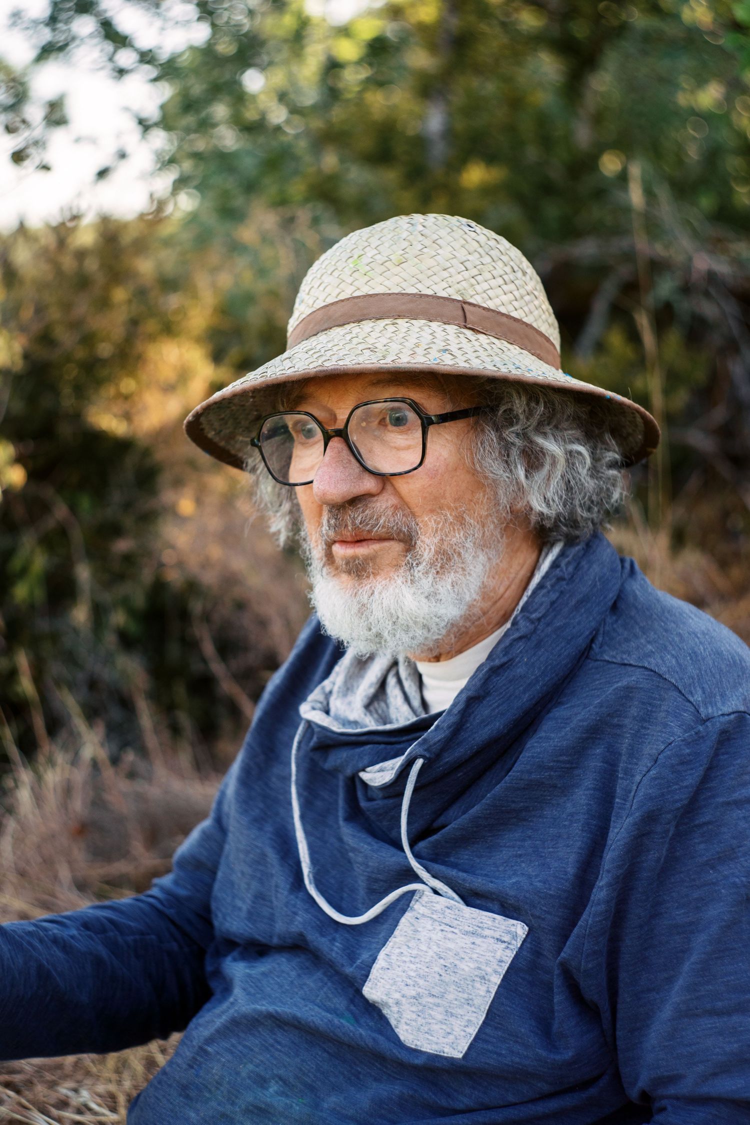 Older bearded man wearing straw hat and glasses sitting outdoors in nature wearing blue sweater.