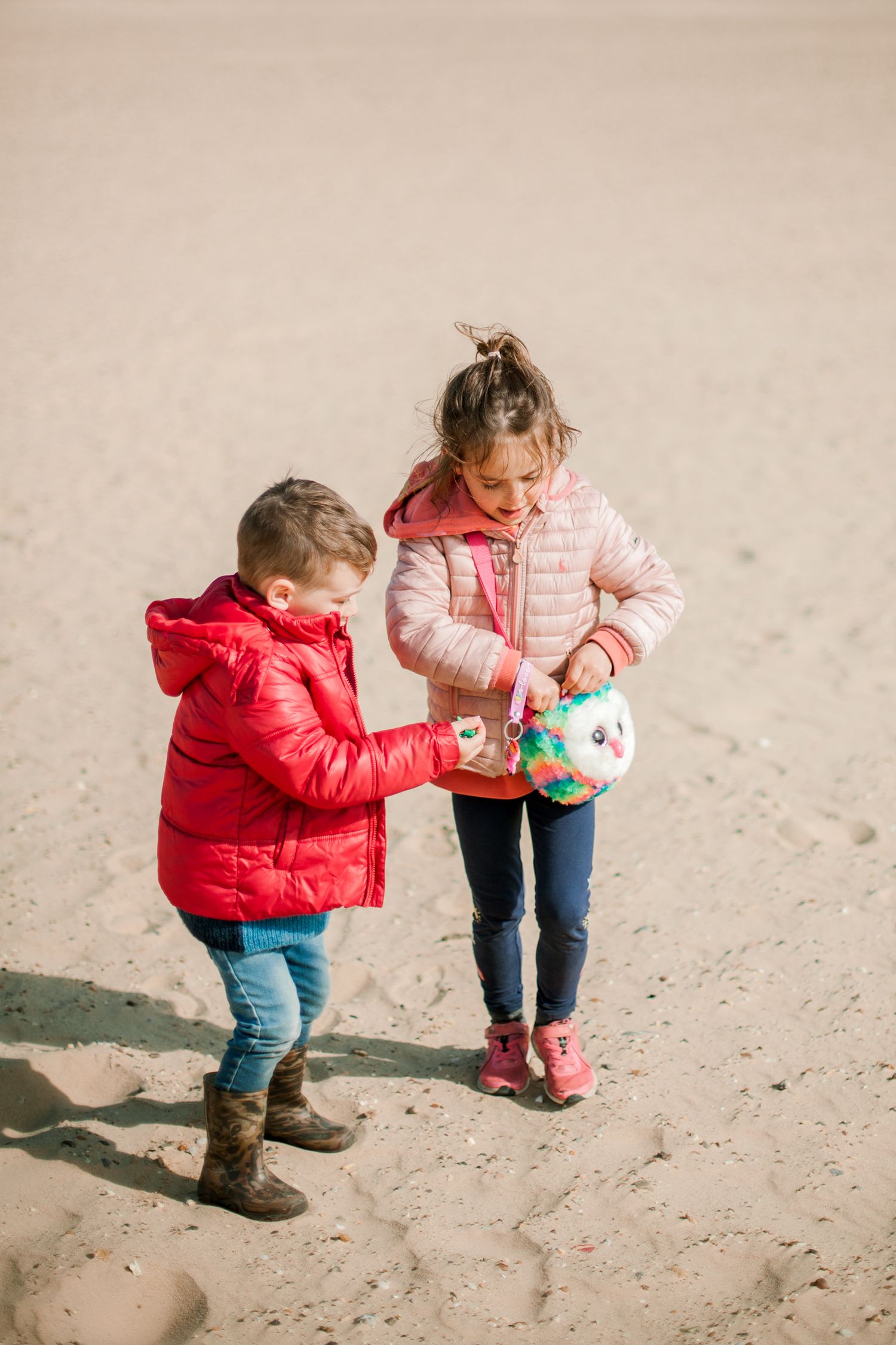 Two young children in colorful winter jackets playing with a toy on a sandy beach on a sunny day.