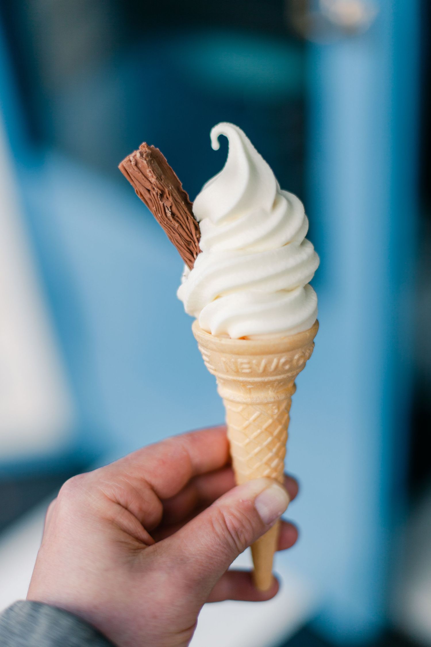 Hand holding a soft serve vanilla ice cream cone with a chocolate flake on a blue background.