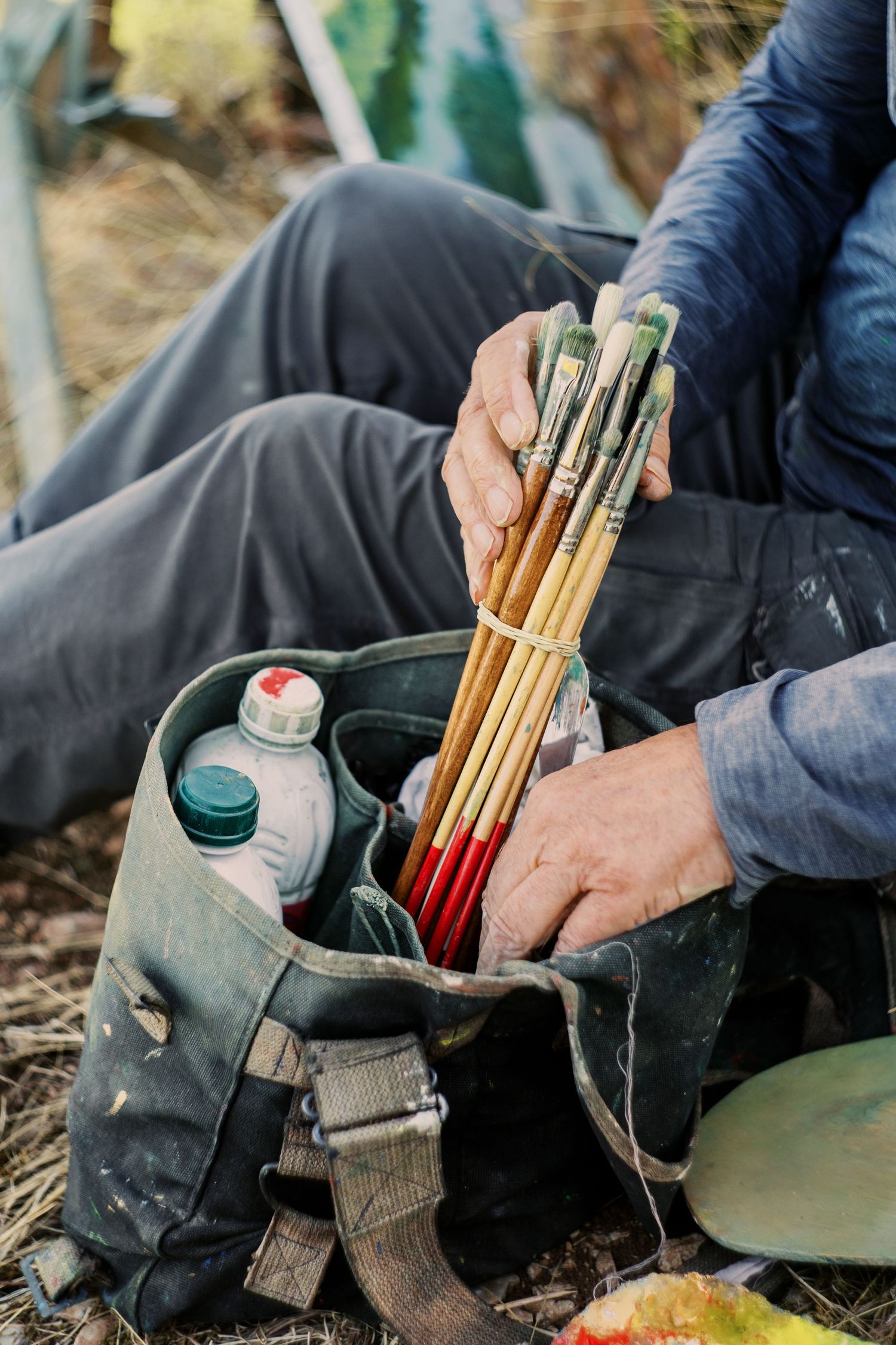 Artist holding paint brushes over a bag with paint supplies outdoors on the ground.