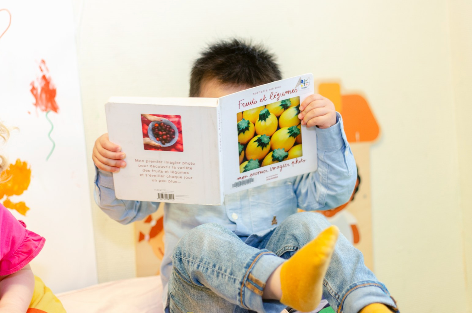 Young boy holds up a colorful fruit book, covering his face while reading in a bright classroom setting.