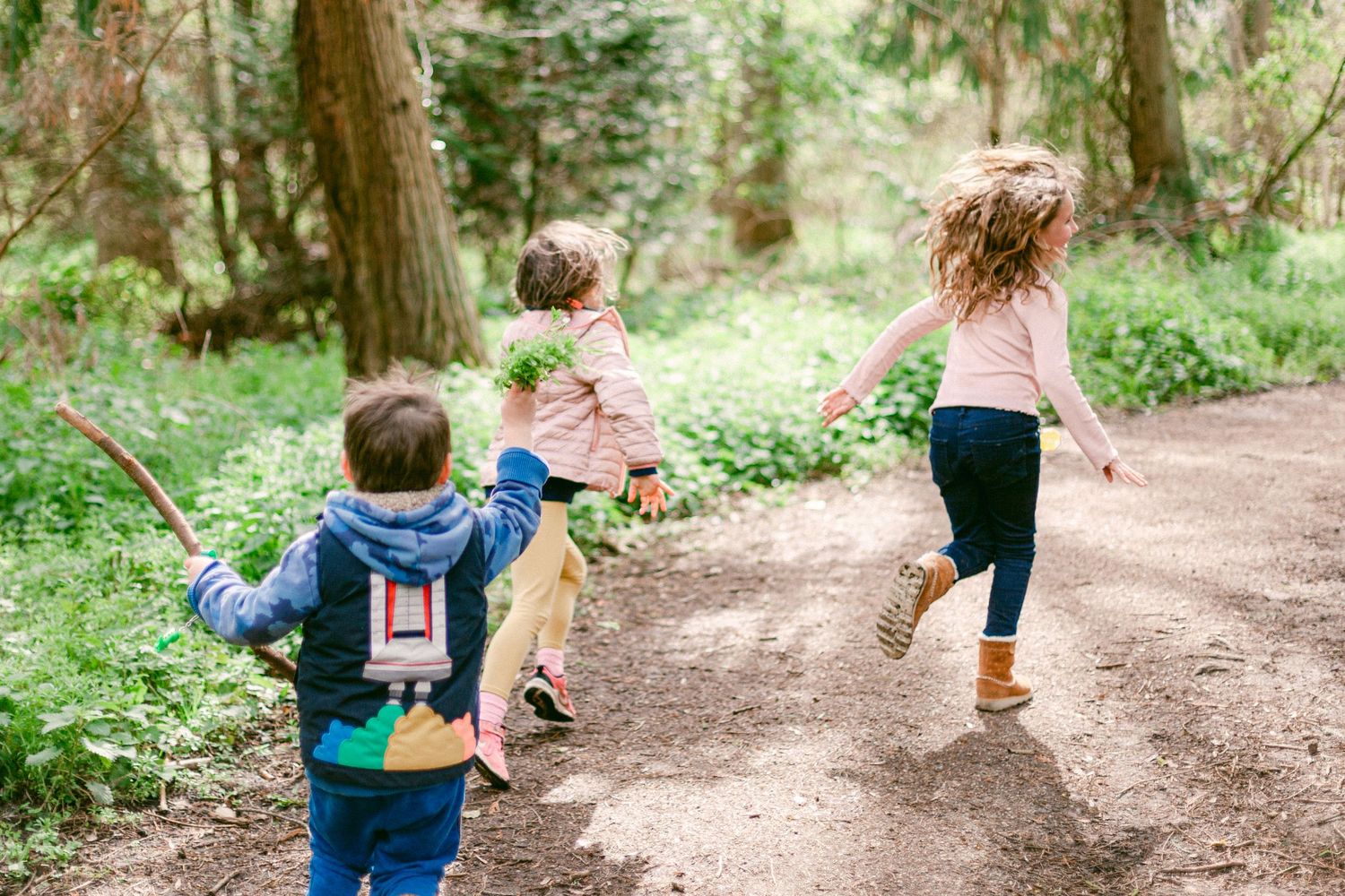 Three children running joyfully along a forest trail surrounded by lush green trees and plants.