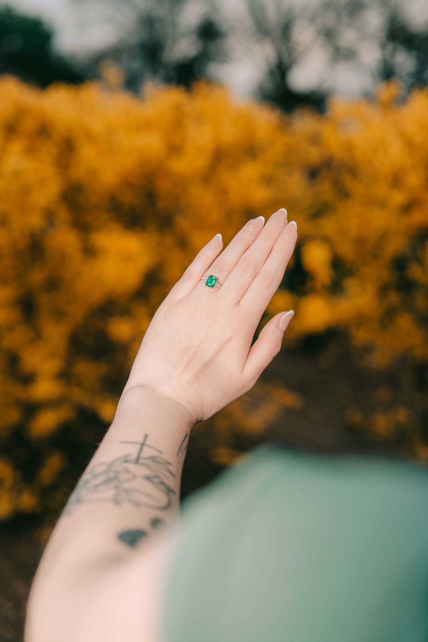Hand wearing an emerald ring stretches upward against a backdrop of golden autumn foliage, with delicate tattoo visible on wrist.
