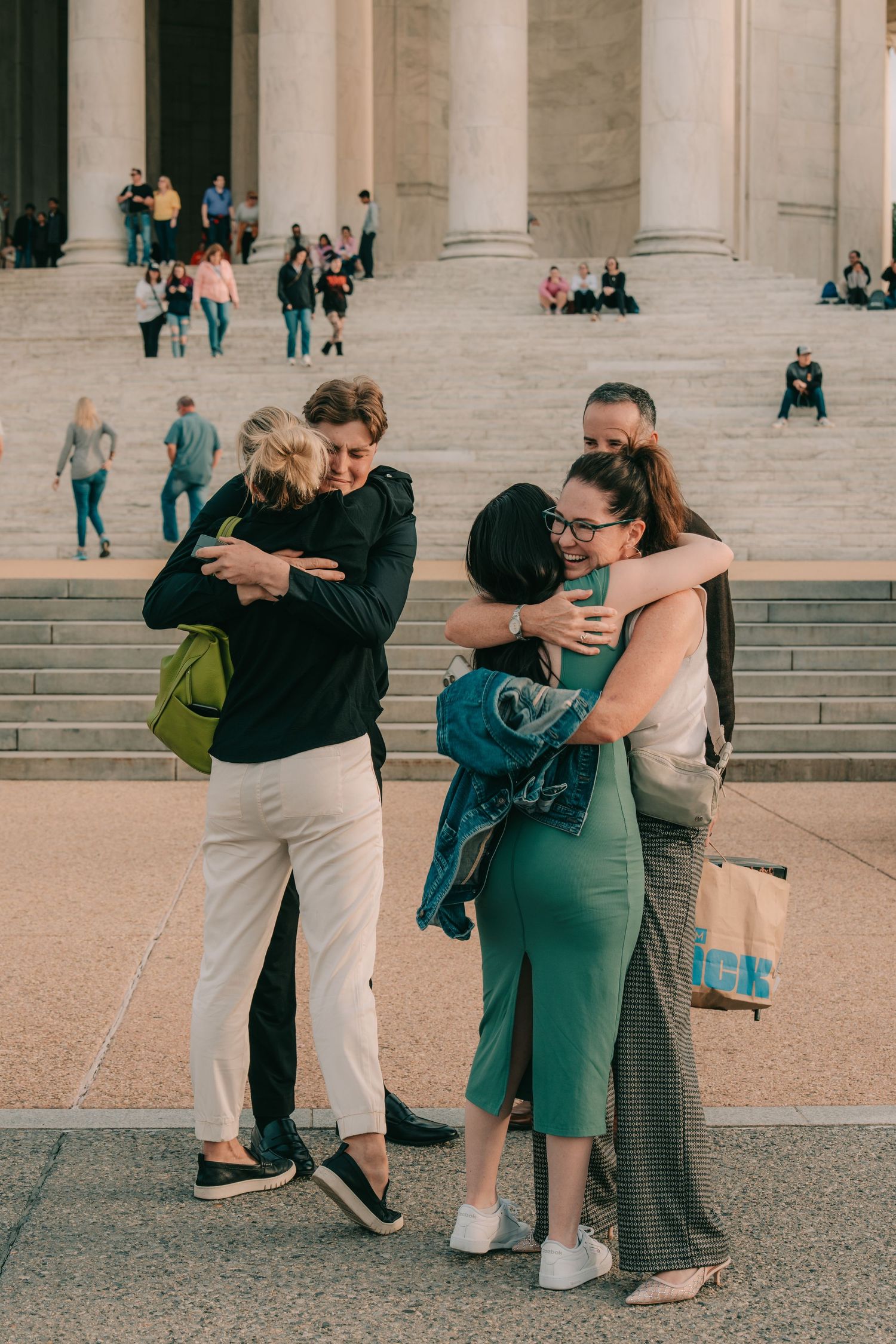 Friends sharing emotional embraces on the steps of a grand building with classical columns in the background.