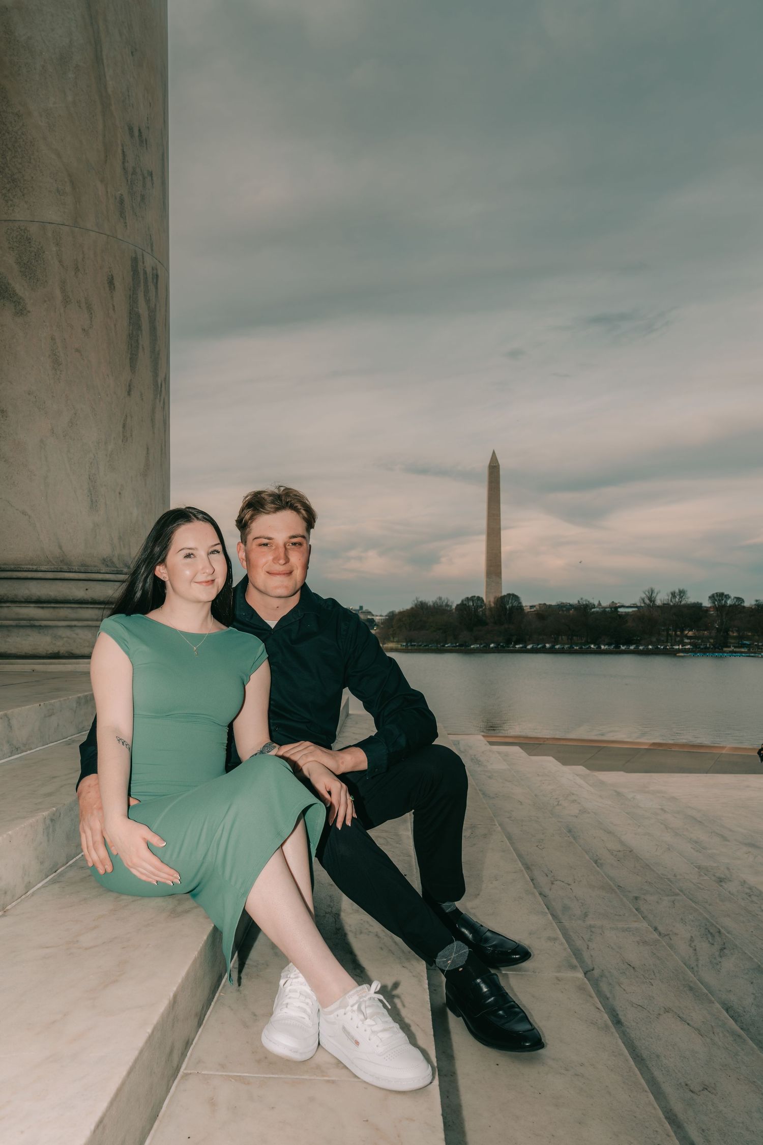 A couple sits on marble steps at the Jefferson Memorial with the Washington Monument visible across the Tidal Basin.