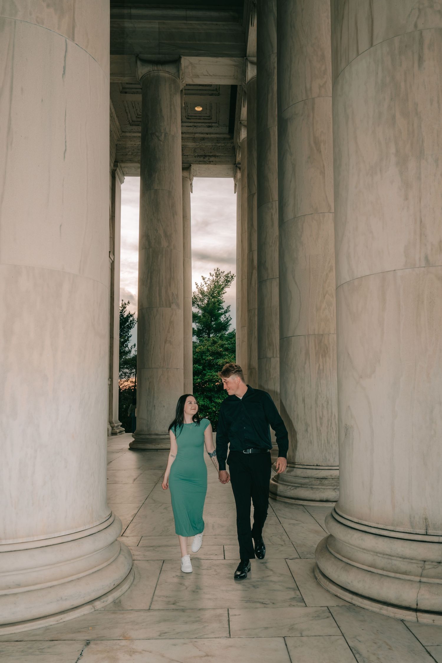 A couple walks between grand classical columns at sunset, with the woman in a teal dress and man in dark clothing.