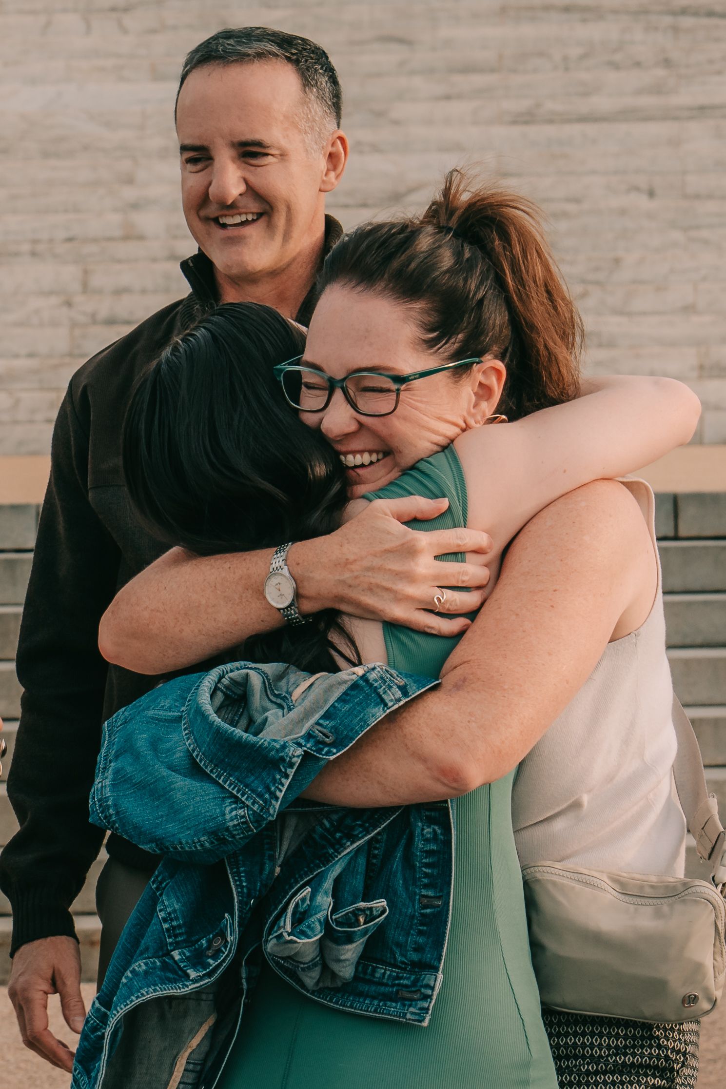 A group sharing a joyful embrace while smiling and laughing together against a neutral wall background.