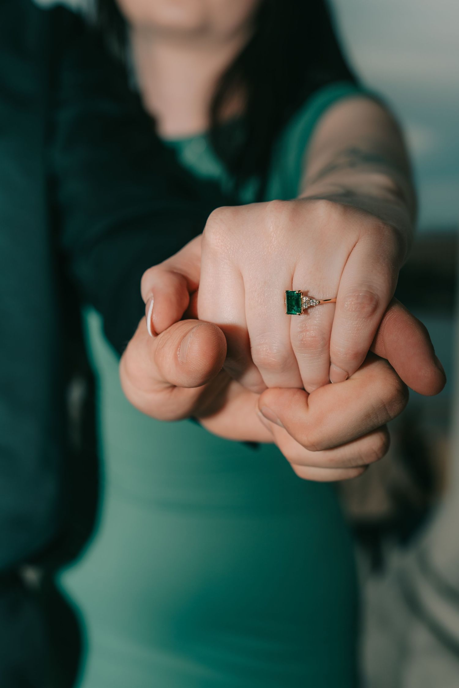 Close-up shot of hands wearing emerald engagement ring against a blurred teal green background.