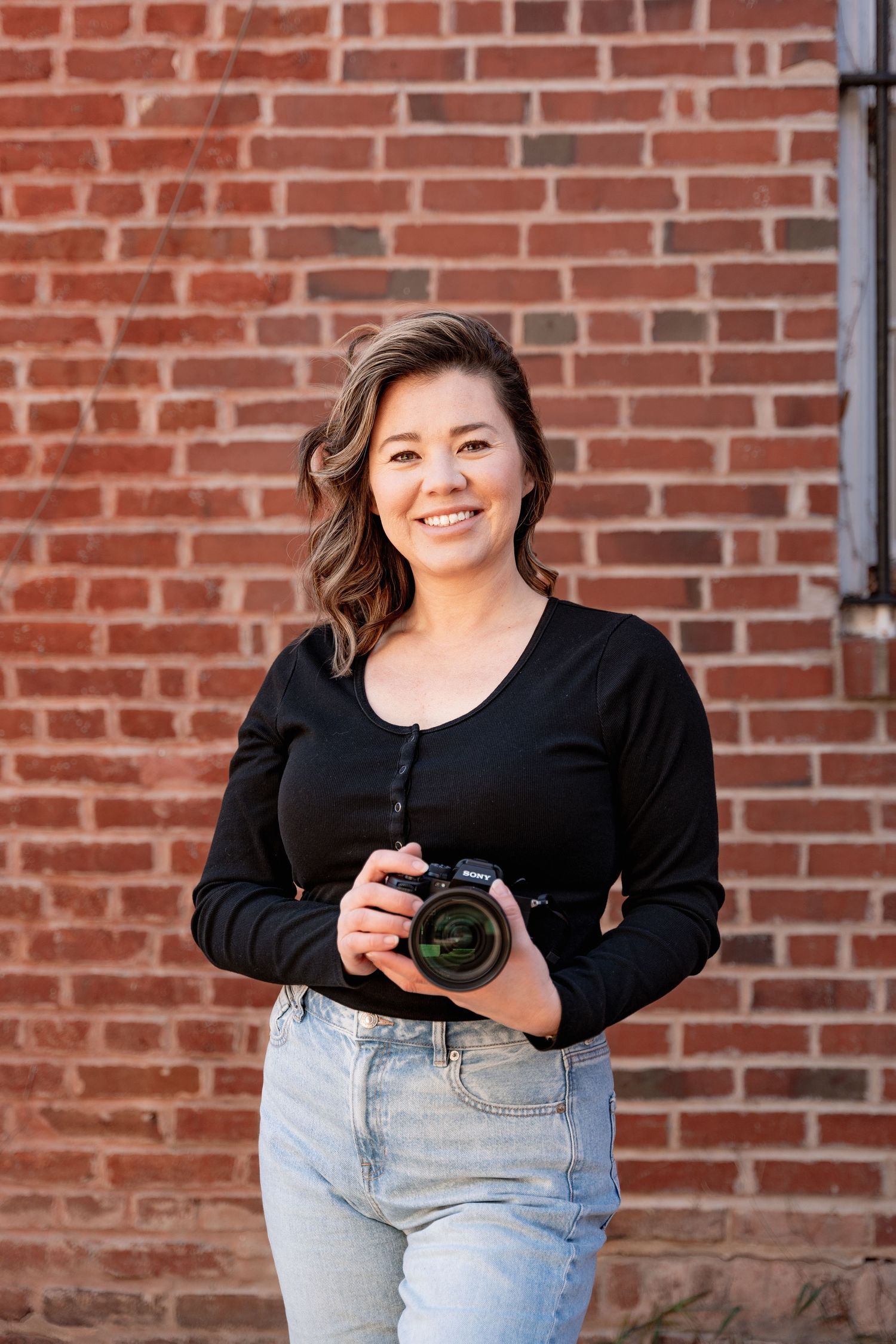 Professional photographer in black top and jeans holding camera against brick wall background.