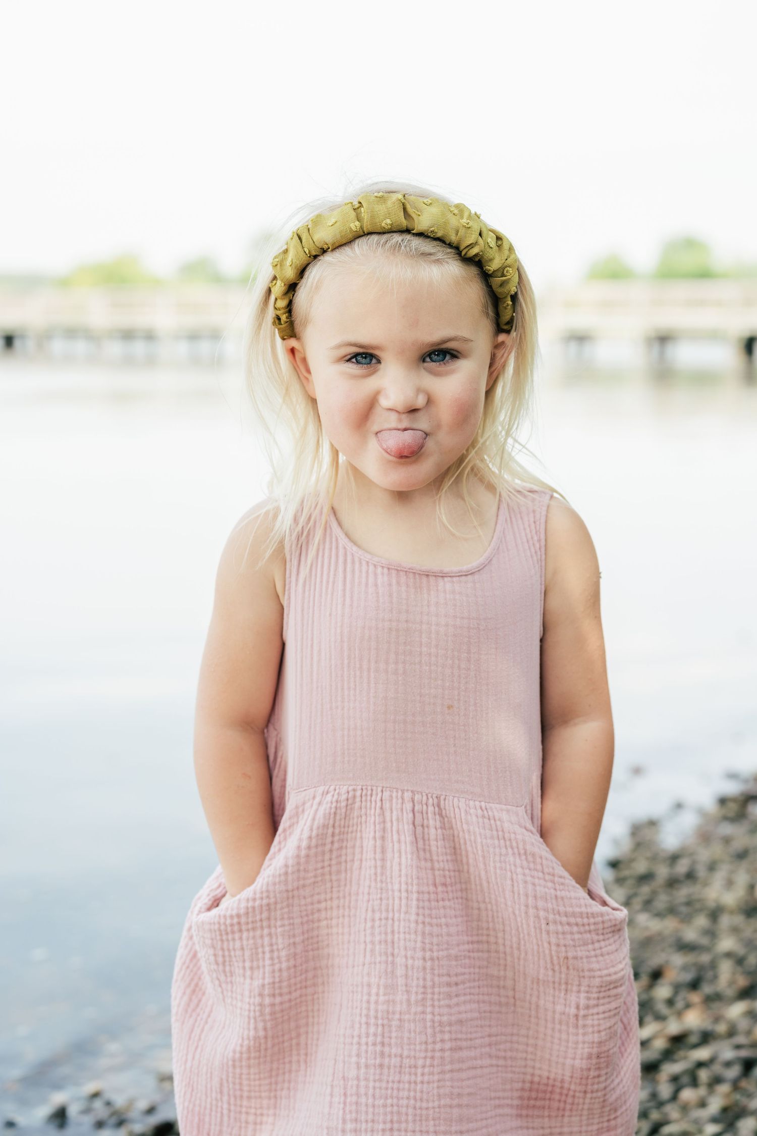 Child in pink sleeveless dress and yellow headband making silly expression by waterfront.