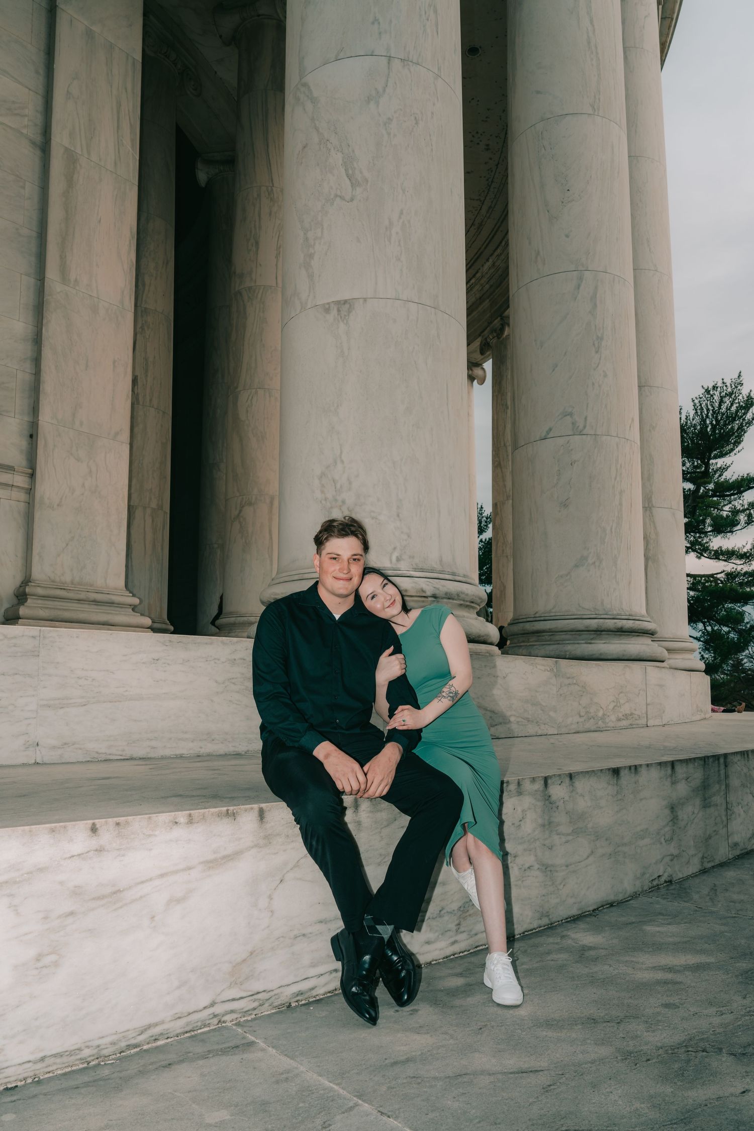 A couple poses together on stone steps between classical white marble columns of a grand building.