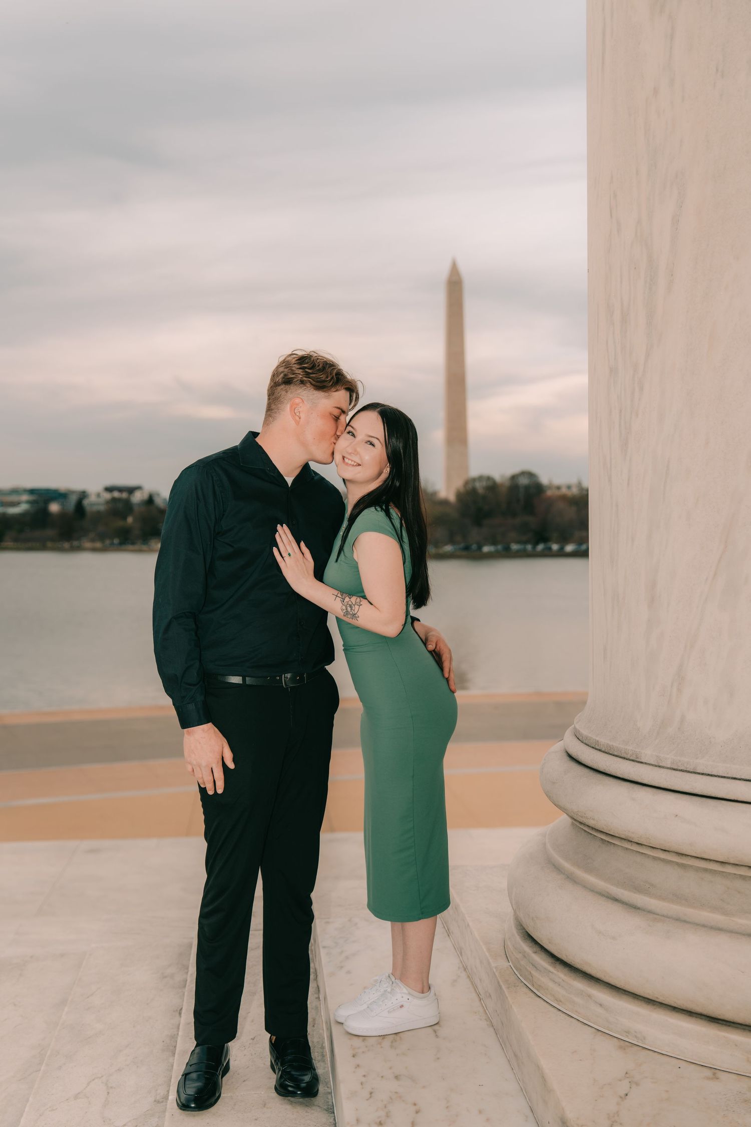 Romantic couple embraces at Jefferson Memorial with Washington Monument visible in background at sunset.