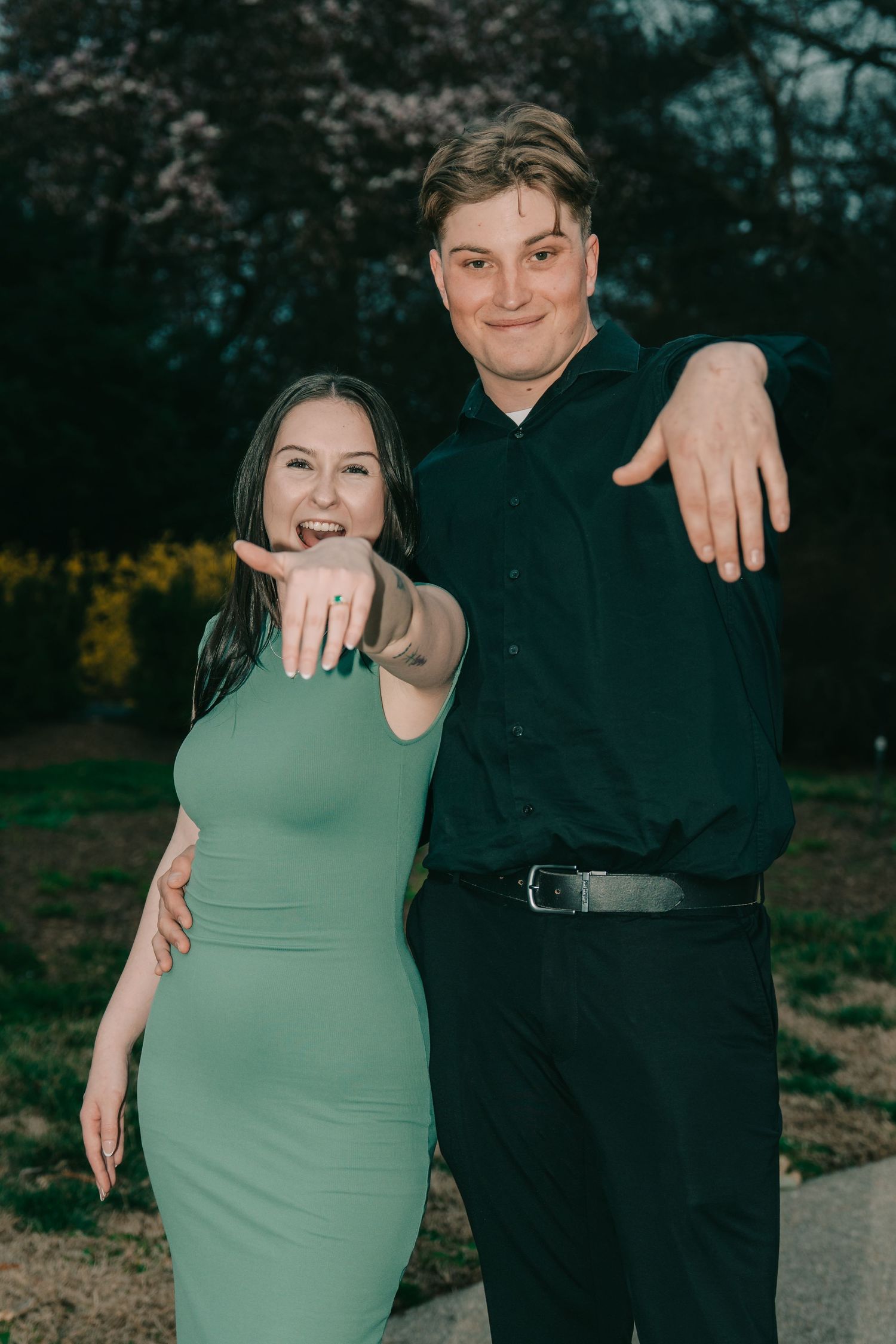A couple poses outdoors showing off their rings, with the woman wearing a sage green dress and the man in a black outfit.
