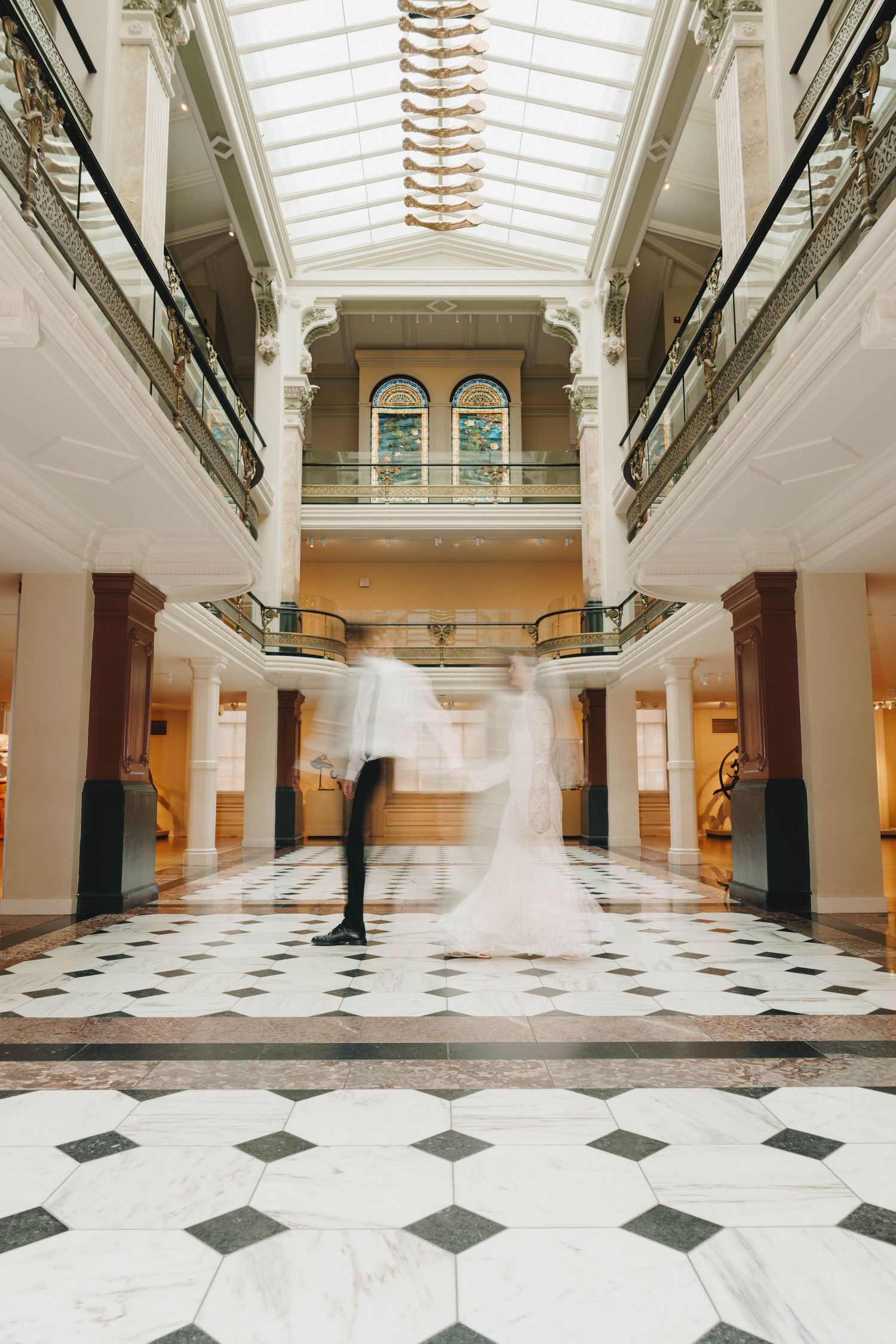 Blurred motion of bride and groom dancing in elegant grand hall with marble floors and glass ceiling.