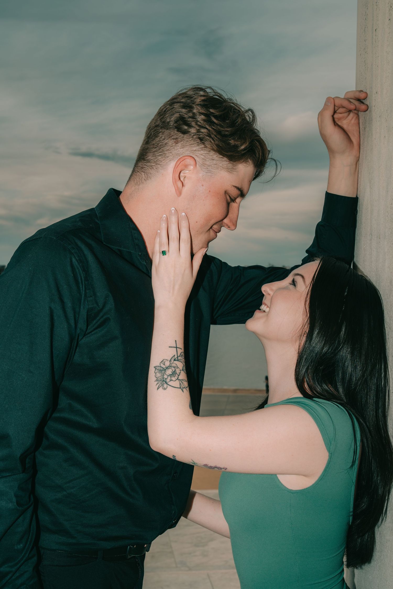 A romantic couple embraces against a cloudy sky backdrop, with one person wearing green and the other in black.