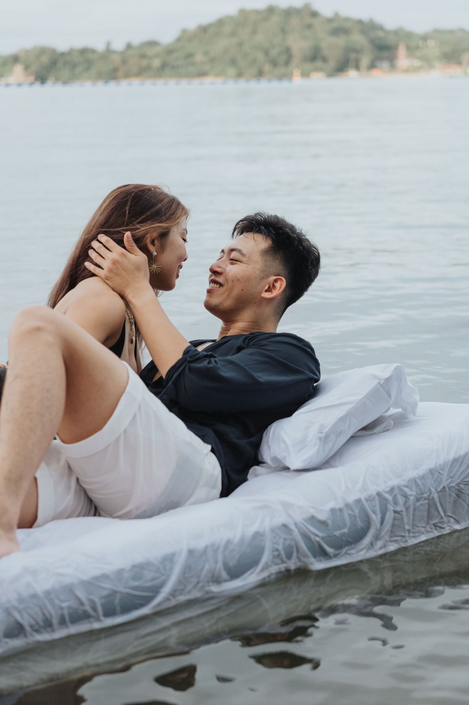 A romantic couple shares an intimate moment on a white mattress by the water.
