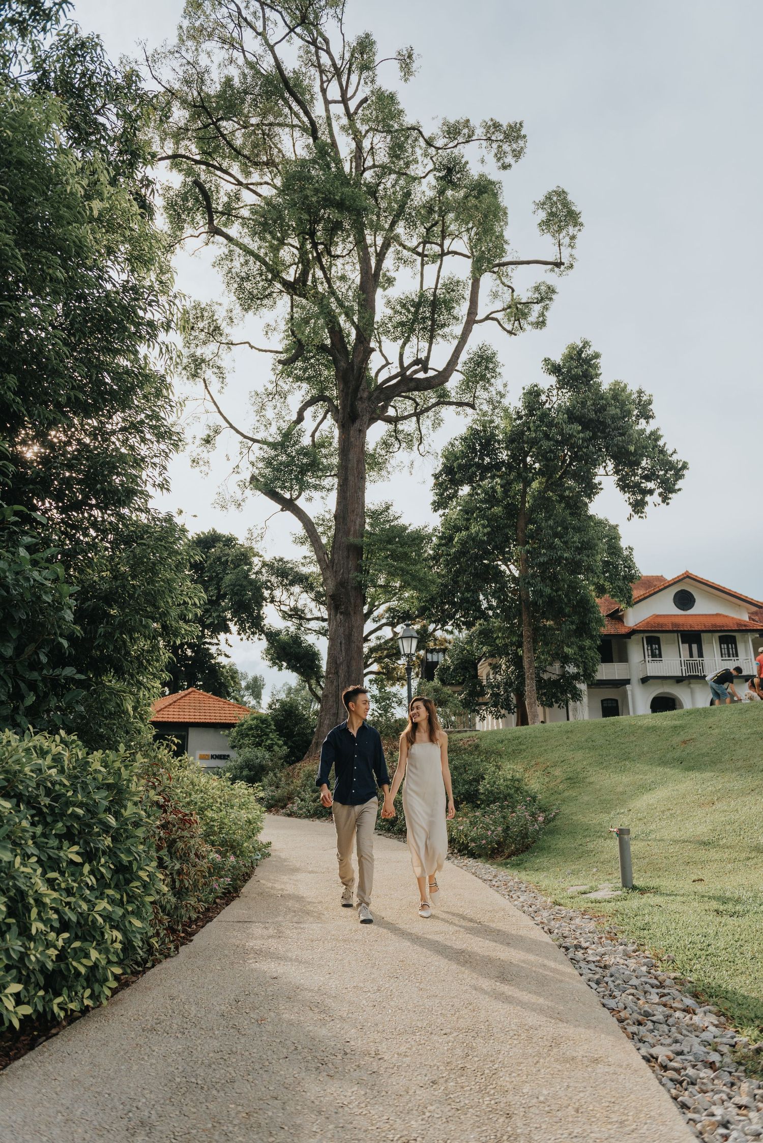 A couple walks along a scenic garden path with tall trees and a colonial-style building in the background.