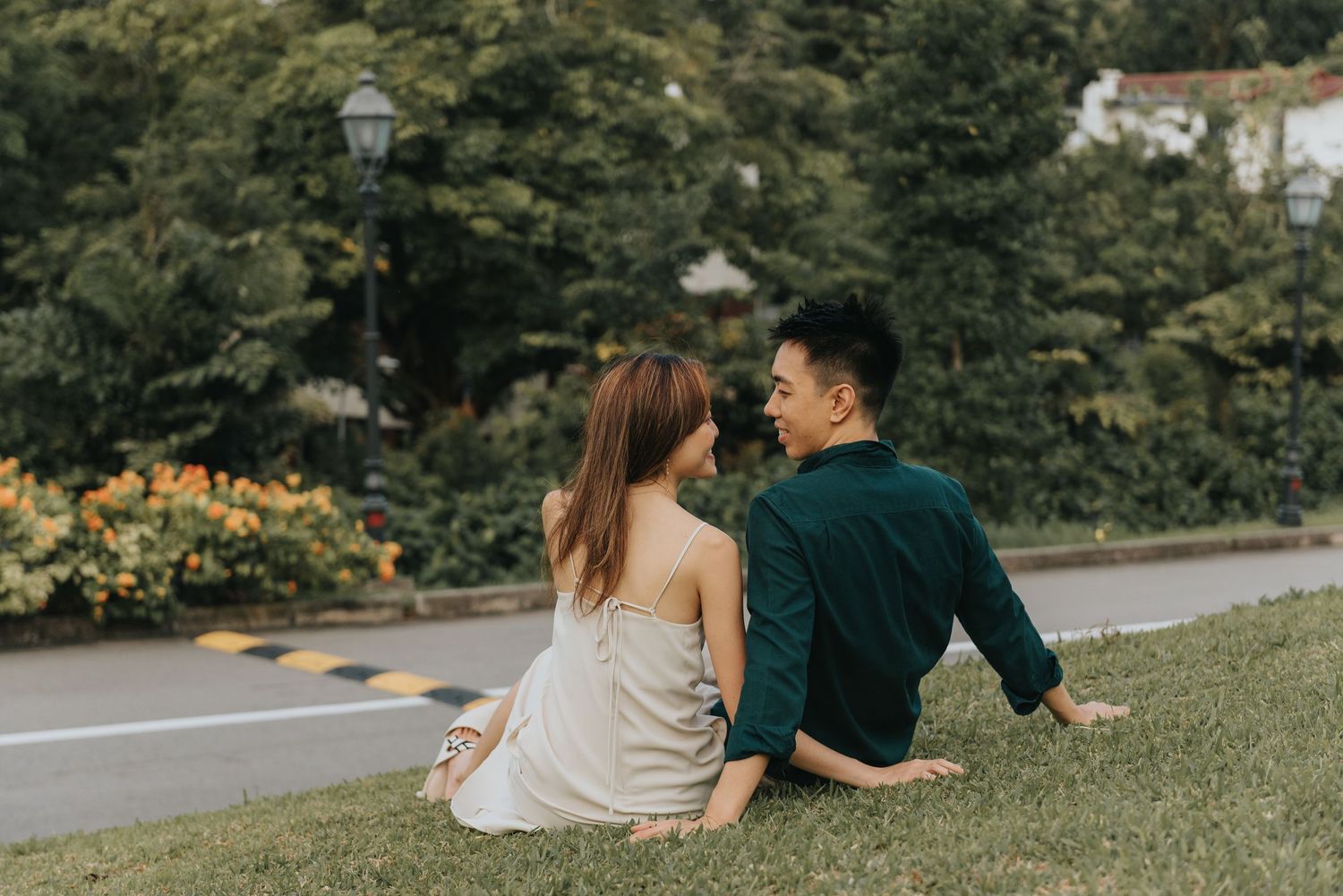 Two people relaxing on grass near path with trees and buildings in background.