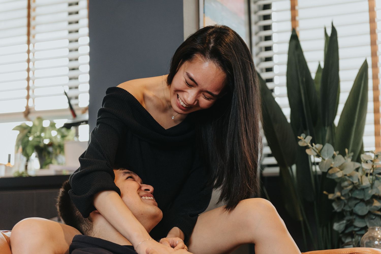 Couple sharing joyful moments on a bed with tropical plants and window blinds in background.