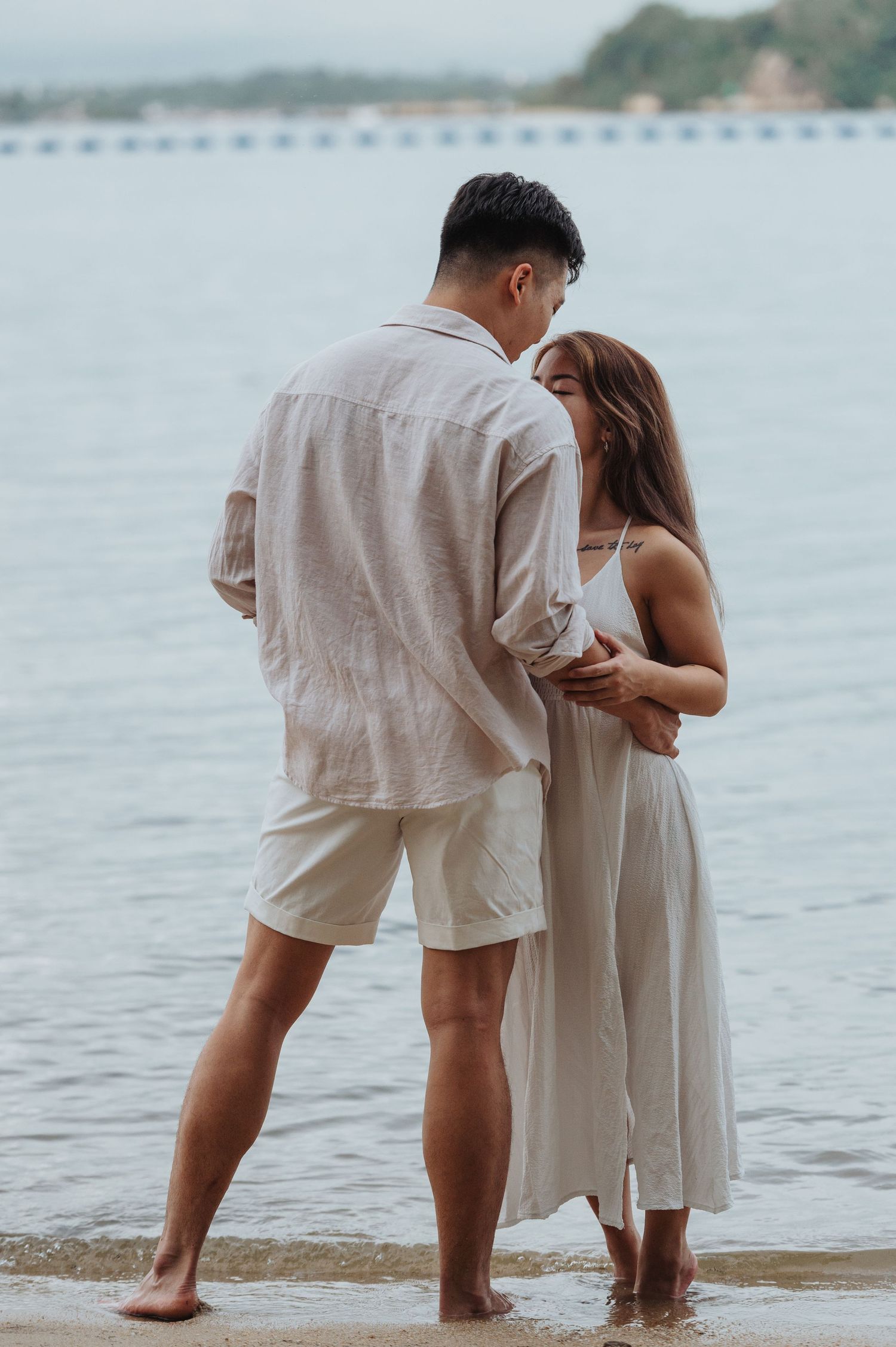 An intimate couple embraces while standing at the edge of calm waters on a tranquil beach.