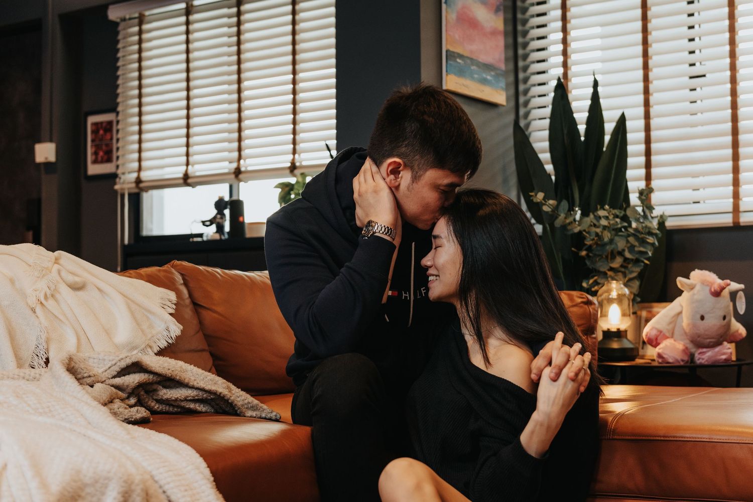 An intimate moment captured on an orange couch with blinds and houseplants in the background.