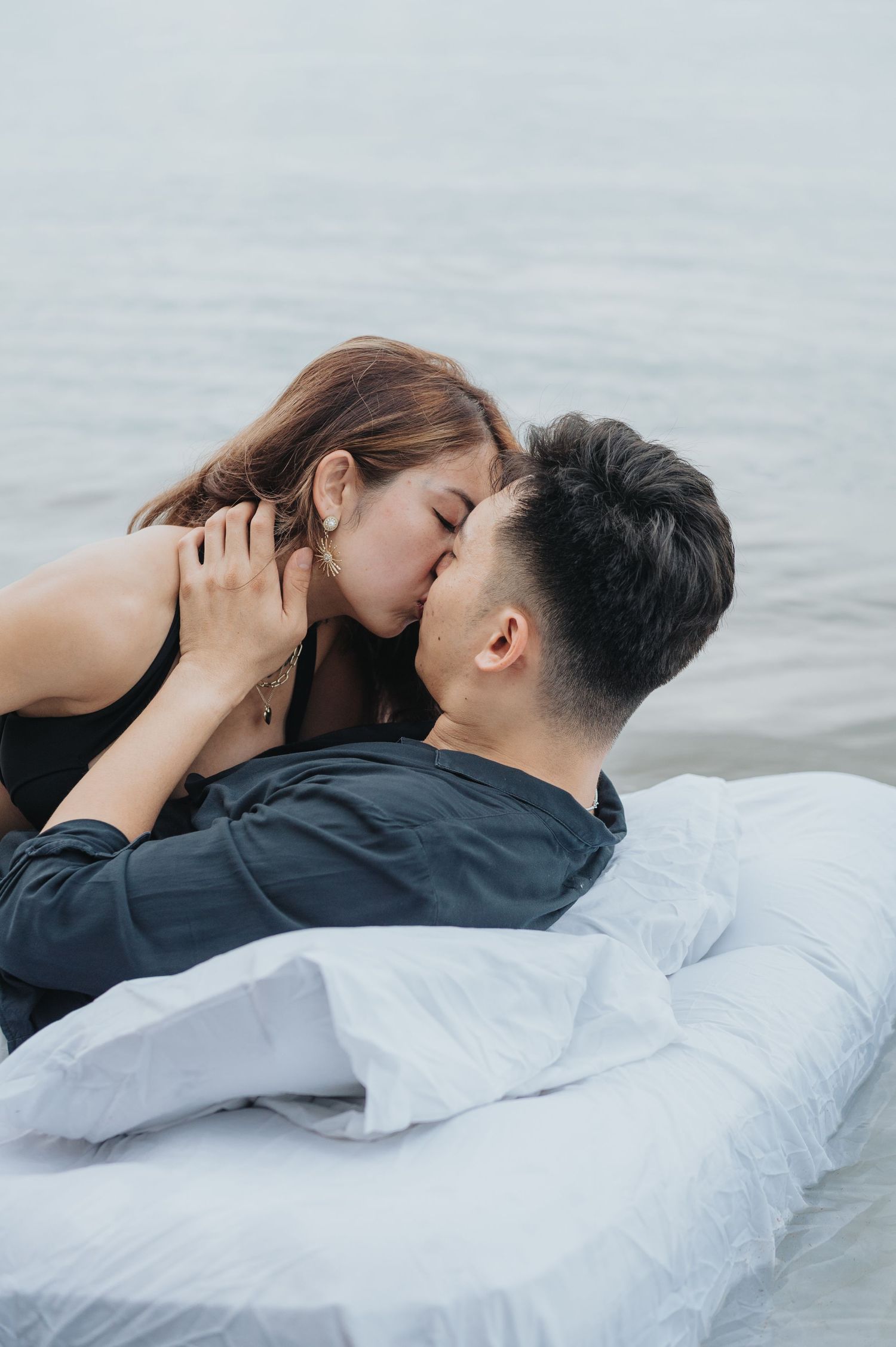 Romantic couple sharing an intimate moment together while laying on white bedding near water.