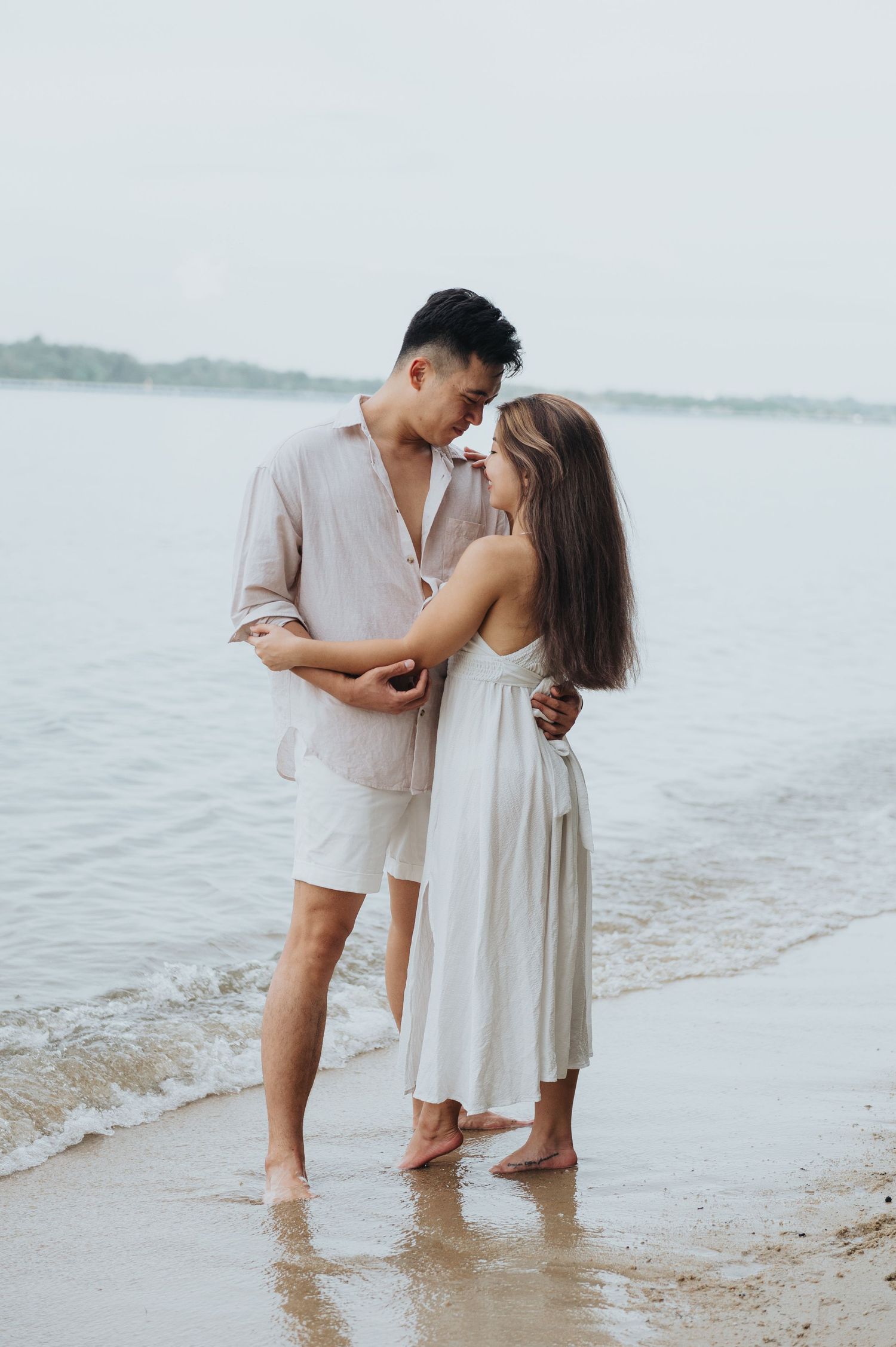 A romantic couple embraces while standing in shallow water at the beach during sunset.