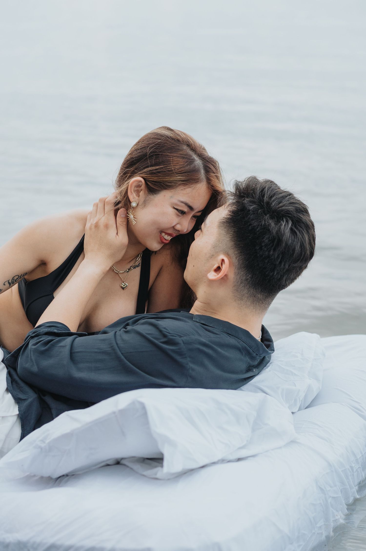 An intimate couple shares laughter while resting on white bedding by the water.