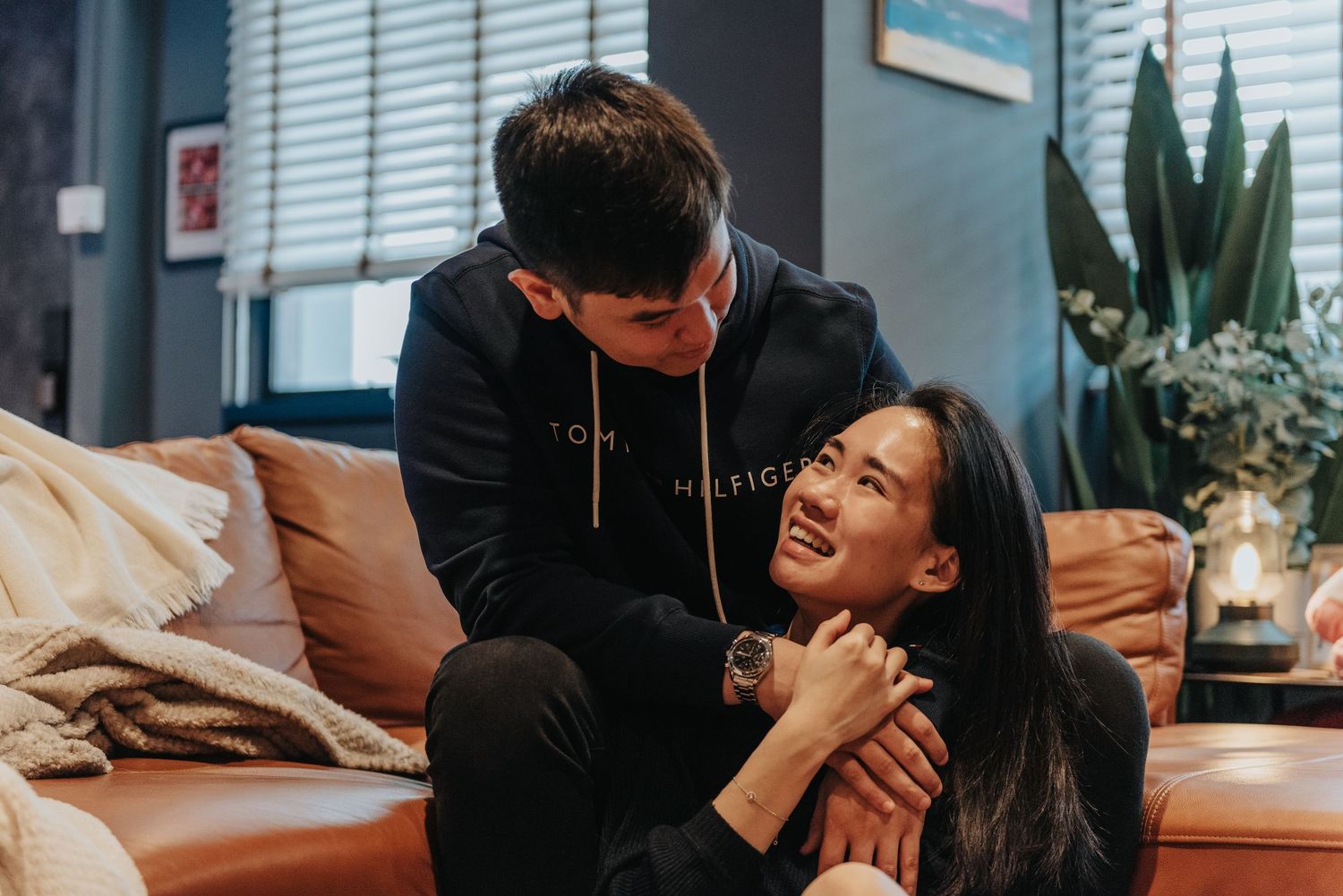 Two people sharing playful laughter together on a tan leather couch with plants in the background.