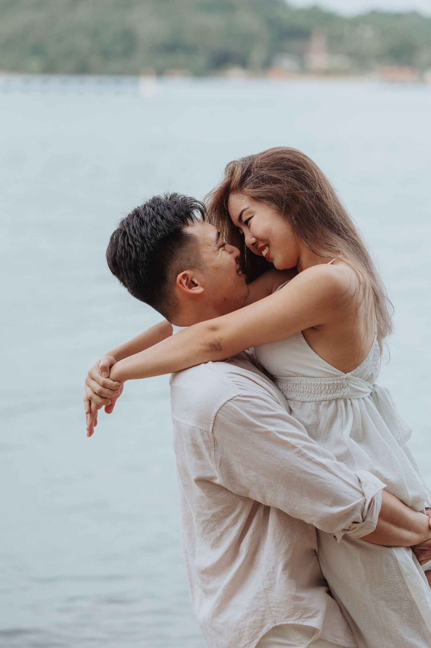 A couple shares an intimate laugh while embracing on a beach with water and hills in background.