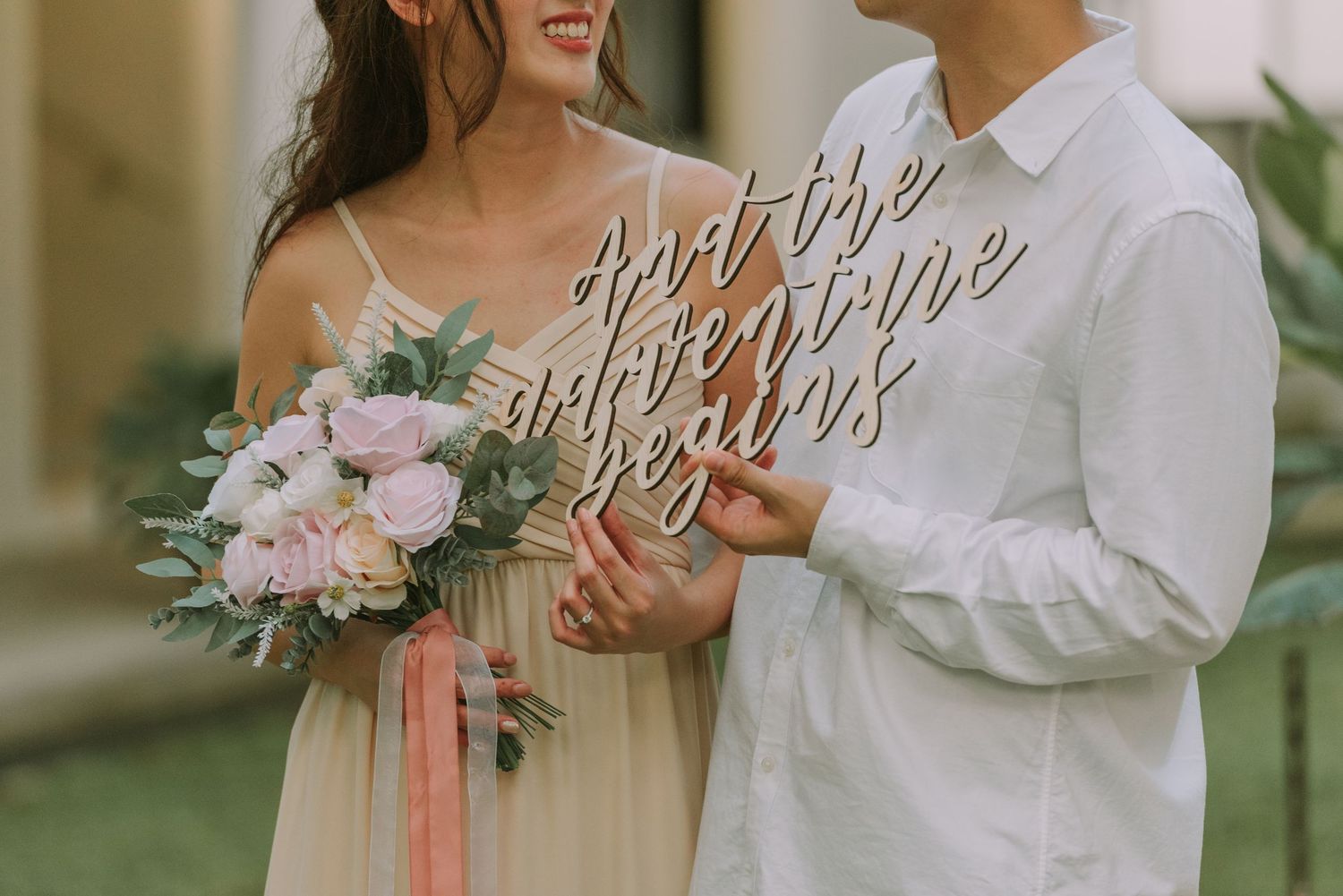 Couple holding decorative signage and pink flower bouquet during engagement photoshoot.