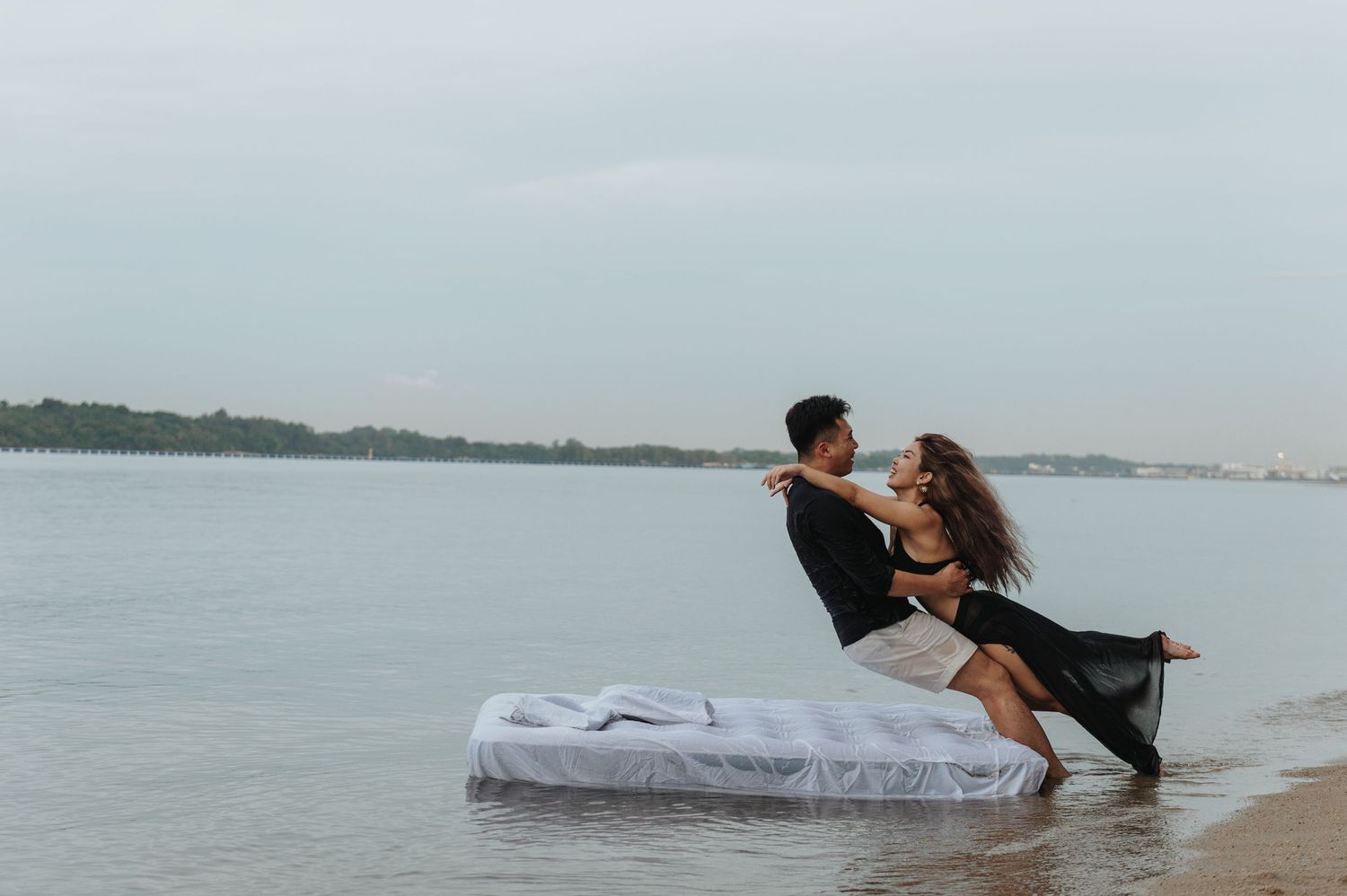 Couple shares tender moment on mattress by ocean shore during cloudy day.