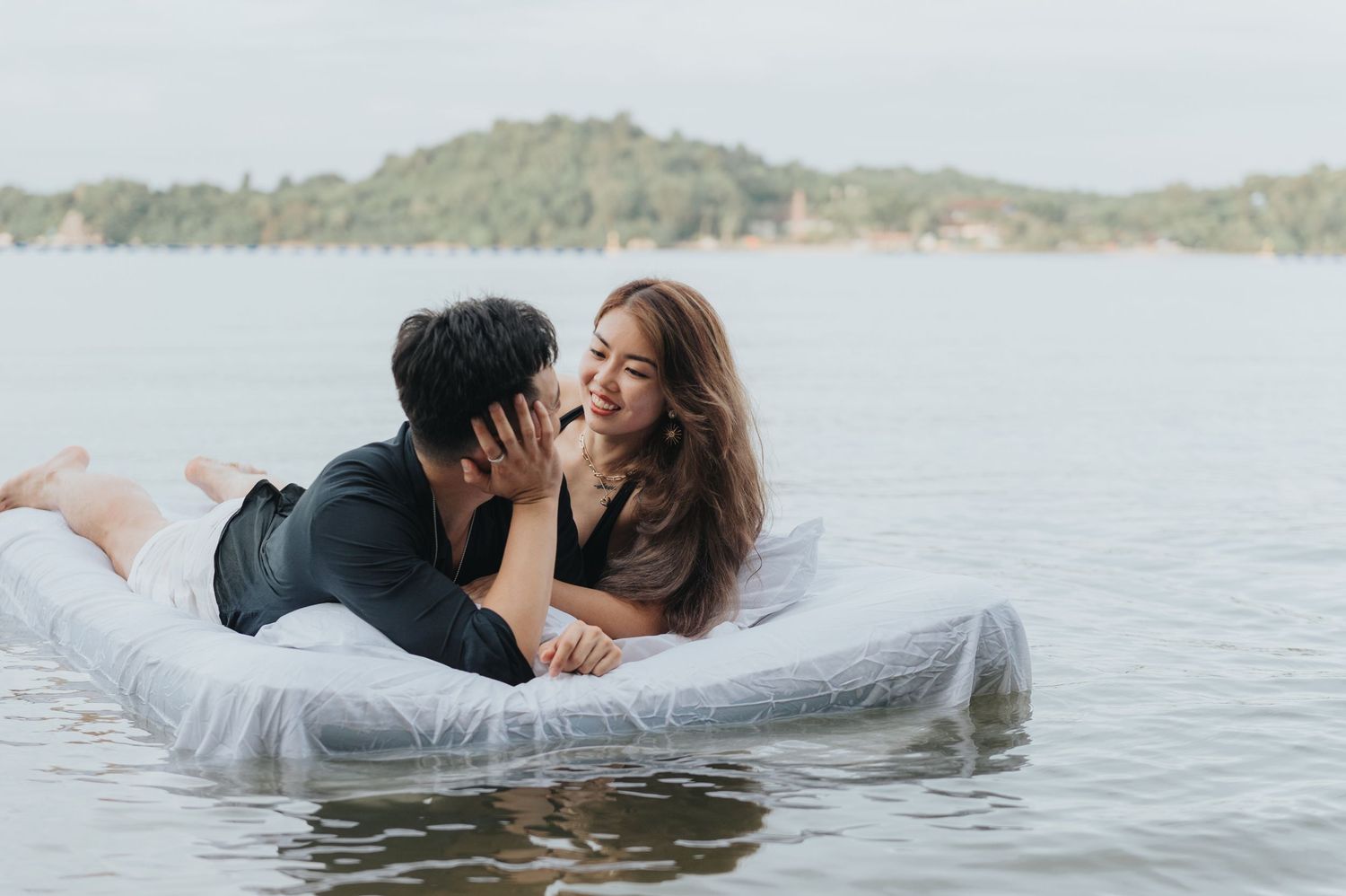 Couple embracing on a floating mattress with serene water and hills in background.