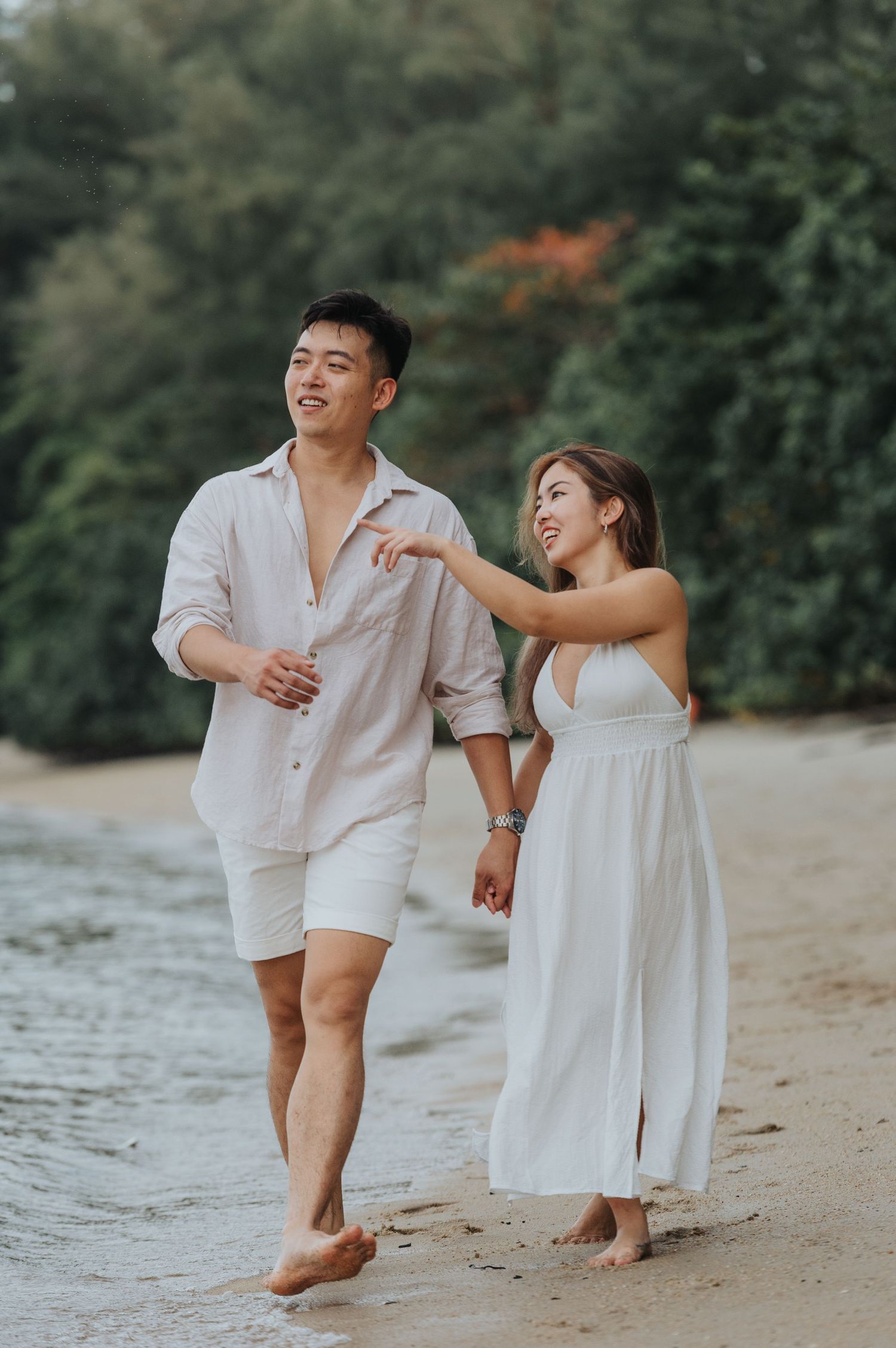 A playful couple laughs while walking barefoot on a scenic beach.